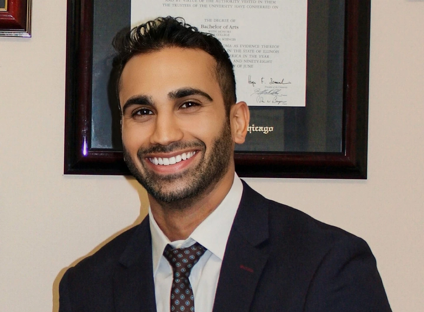 Dr. Chris Devulapalli in dark suit and patterned tie, smiling in front of framed diploma