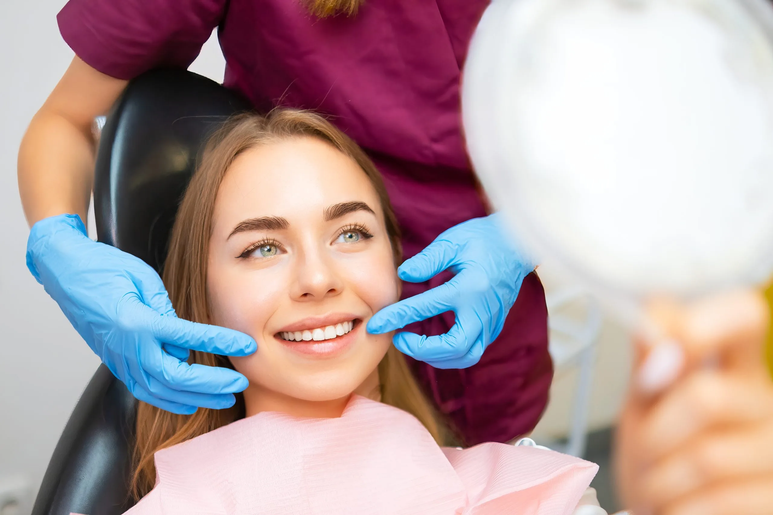 Dentist in blue gloves examining smiling patient's teeth in dental chair