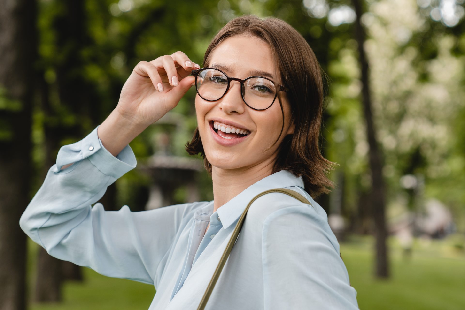 Smiling woman adjusting eyeglasses outdoors in a park setting