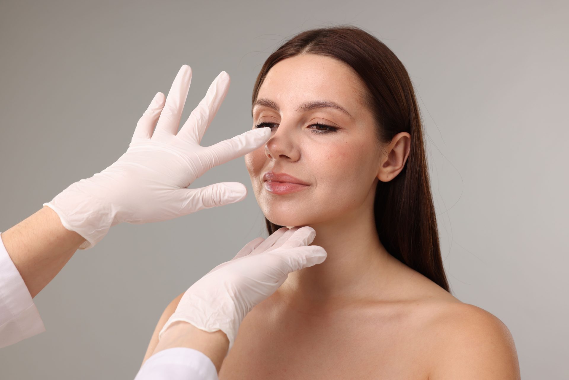 Gloved hands examining a woman's nose and facial structure against a neutral background