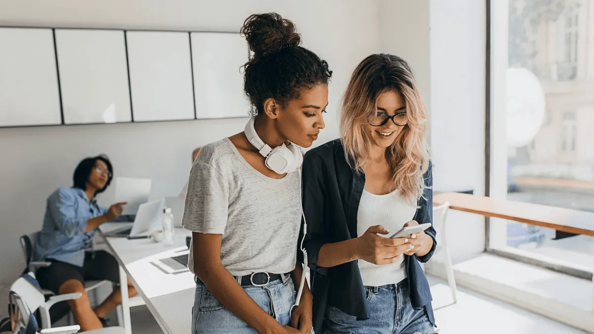 Duas mulheres em um escritório moderno, uma de camiseta cinza e outra com blazer, usam fones de ouvido e analisam informações no celular enquanto sorriem e trabalham em equipe