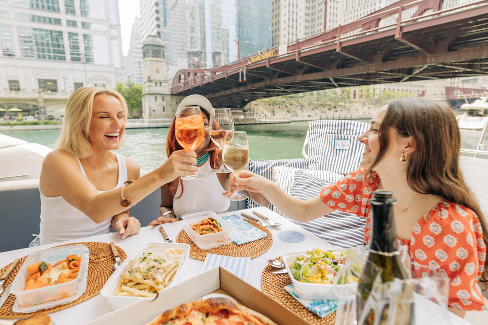 Three women on a boat cheersing, enjoying salad, truffle fries, and pastas