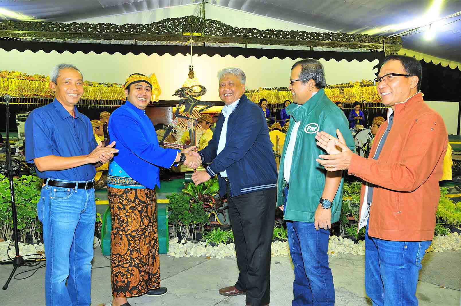  A group of five men are posing for a photo in front of a stage with a traditional Bugis puppet show.