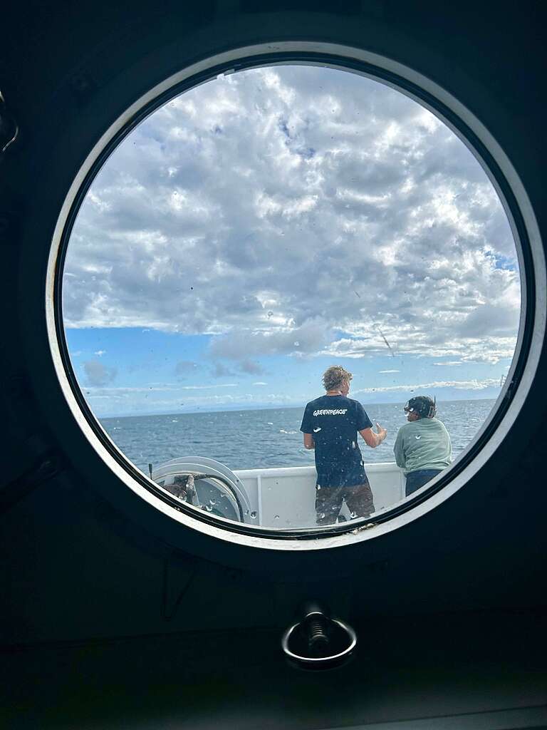 Photo taken through round porthole window looking out to deck where two people are talking with the ocean and sky in the background