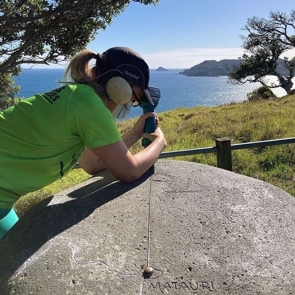 A person in green tshirt and ear protection is using a tool on a smooth rock. The words MATARUI is etched in the rock. A beautiful blue sea and sky is behind them.