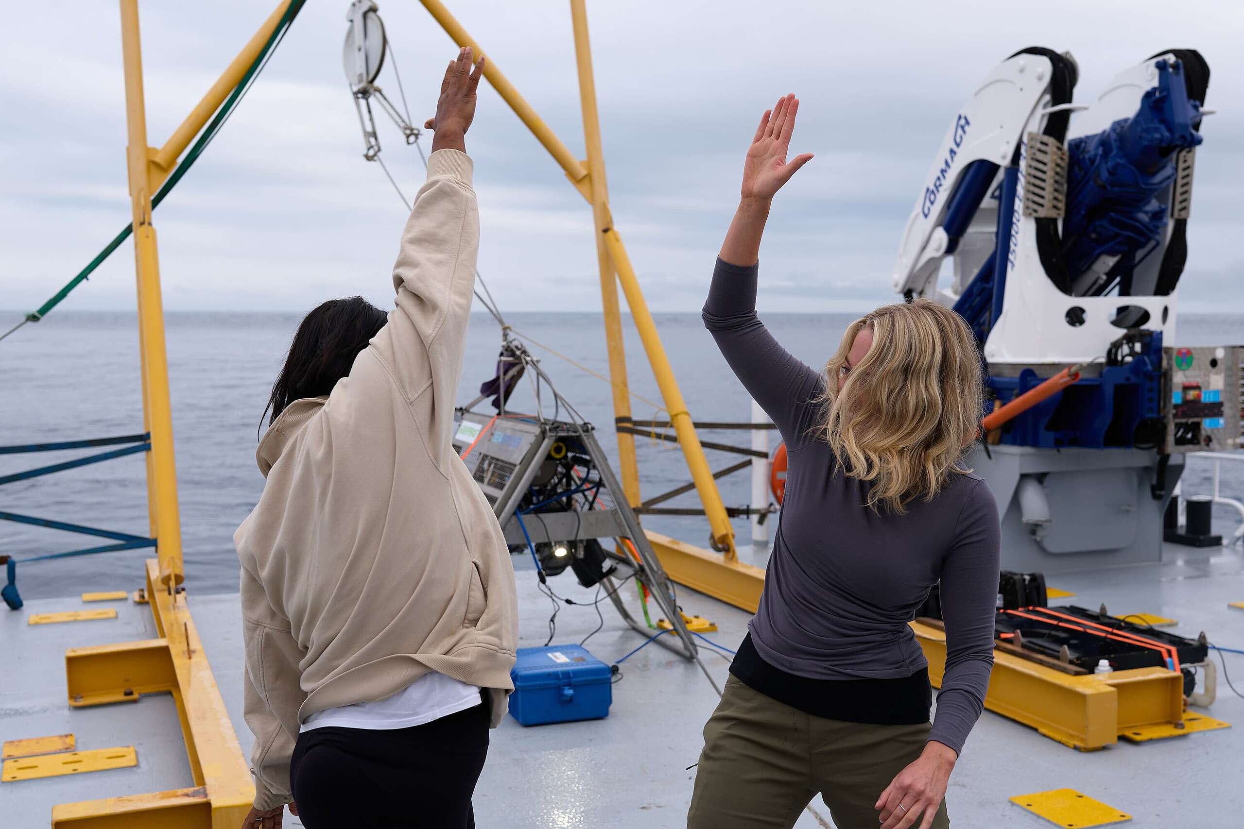 Lead scientist Kat Goddard stands on the left, right arm raised ready to high five Ellie Hooper, expedition lead, to her left. Both look happy.