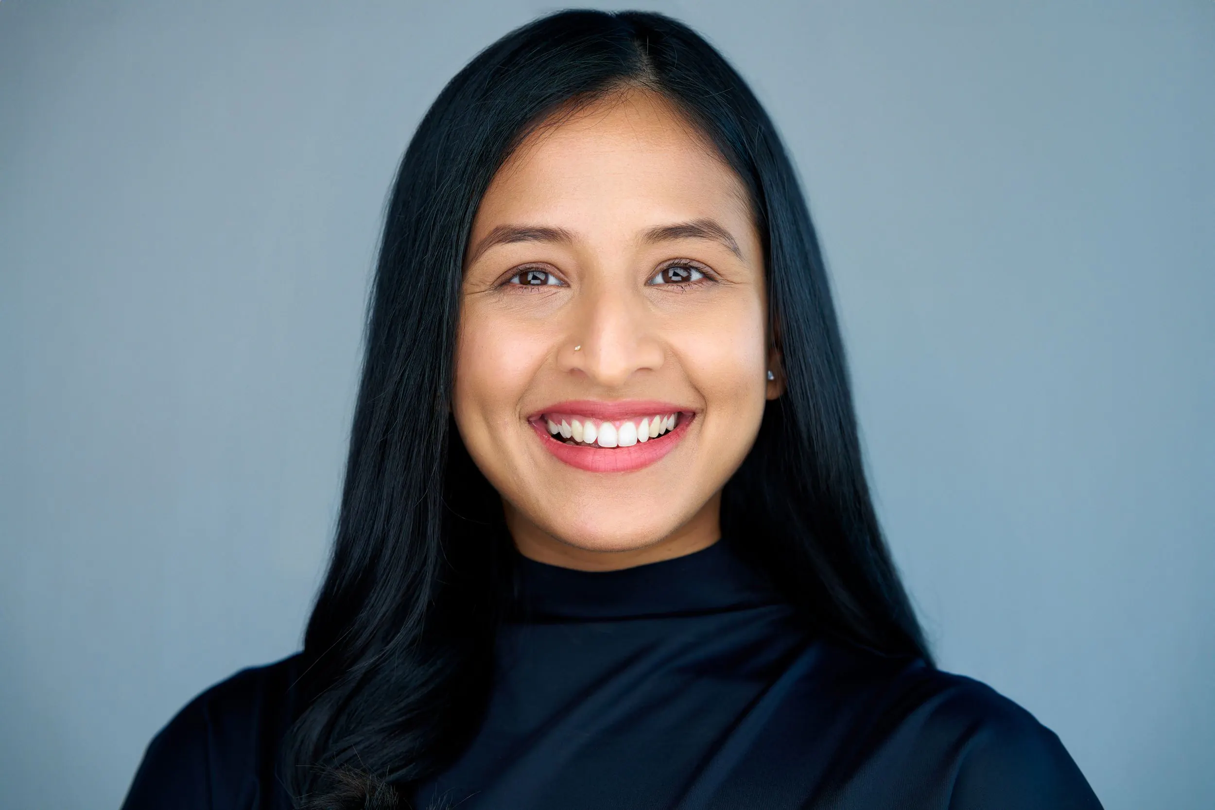 Professional headshot of a smiling person with long dark hair in a black top against a blue backdrop — Shots by Dima, San Jose.