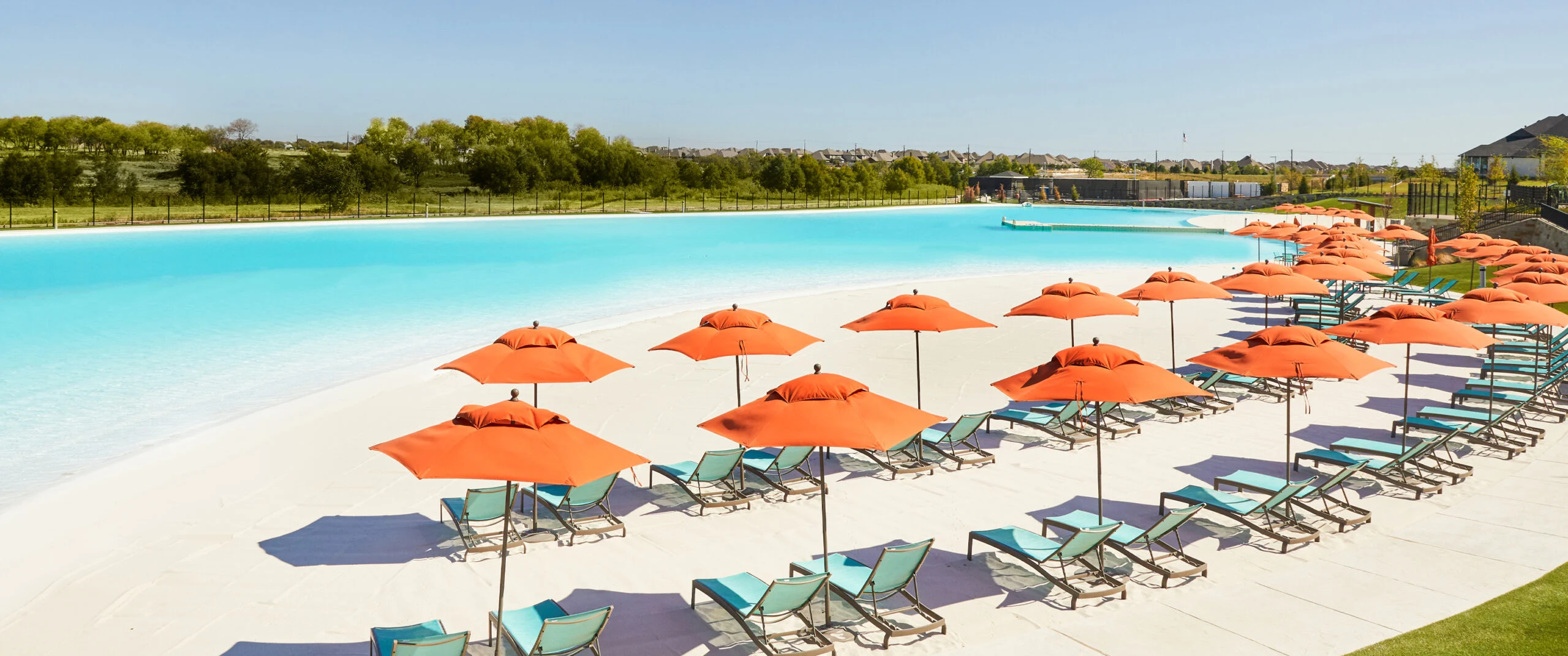 THE LAGOON at Windsong Ranch in Prosper, Texas, with clear blue water, a white sand beach, orange umbrellas, loungers, and homes in the distance