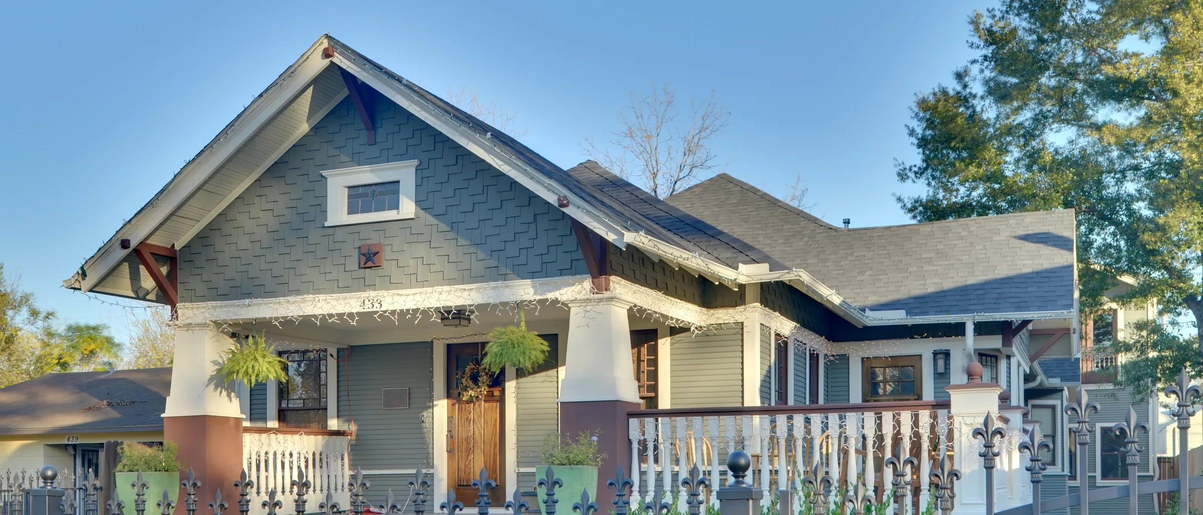 Gray-green shingled Craftsman bungalow with tapered porch columns behind a wrought-iron fence in Houston Heights
