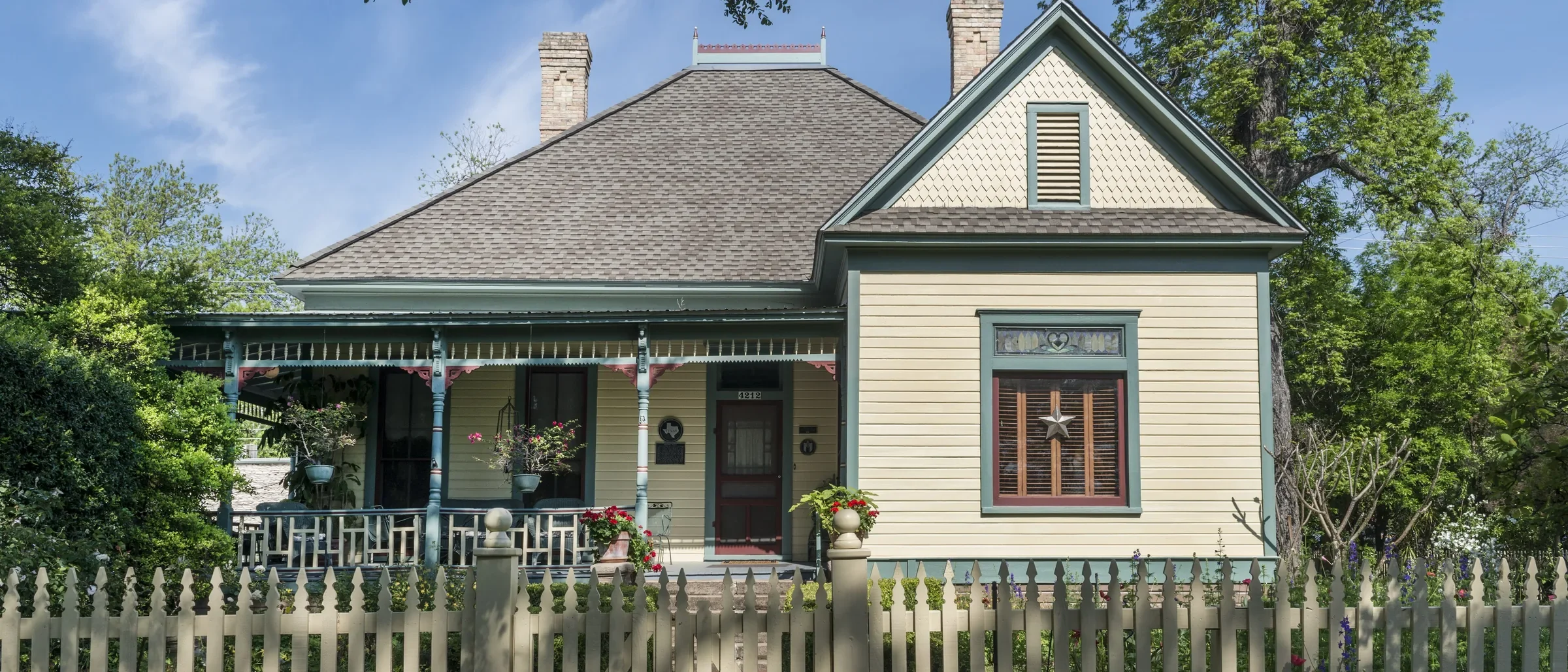 Cream Victorian cottage with teal trim, a wraparound porch, and a Texas star on a shuttered window behind a picket fence in Austin's Hyde Park neighborhood
