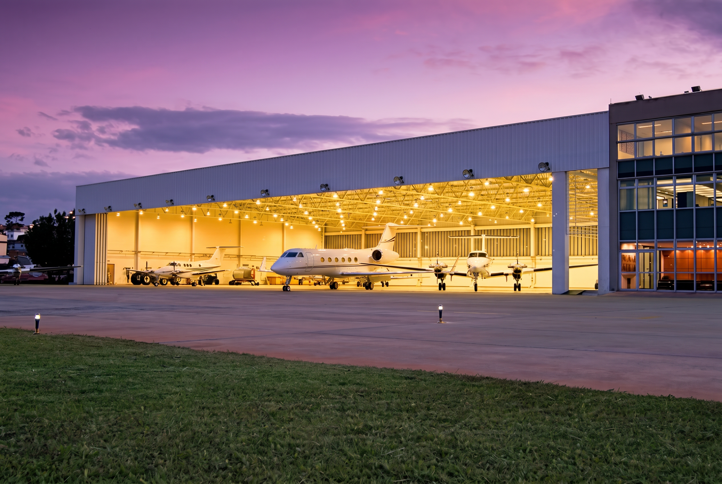 Private jets parked in a warmly lit FBO hangar at dusk under a pink and lavender sunset sky