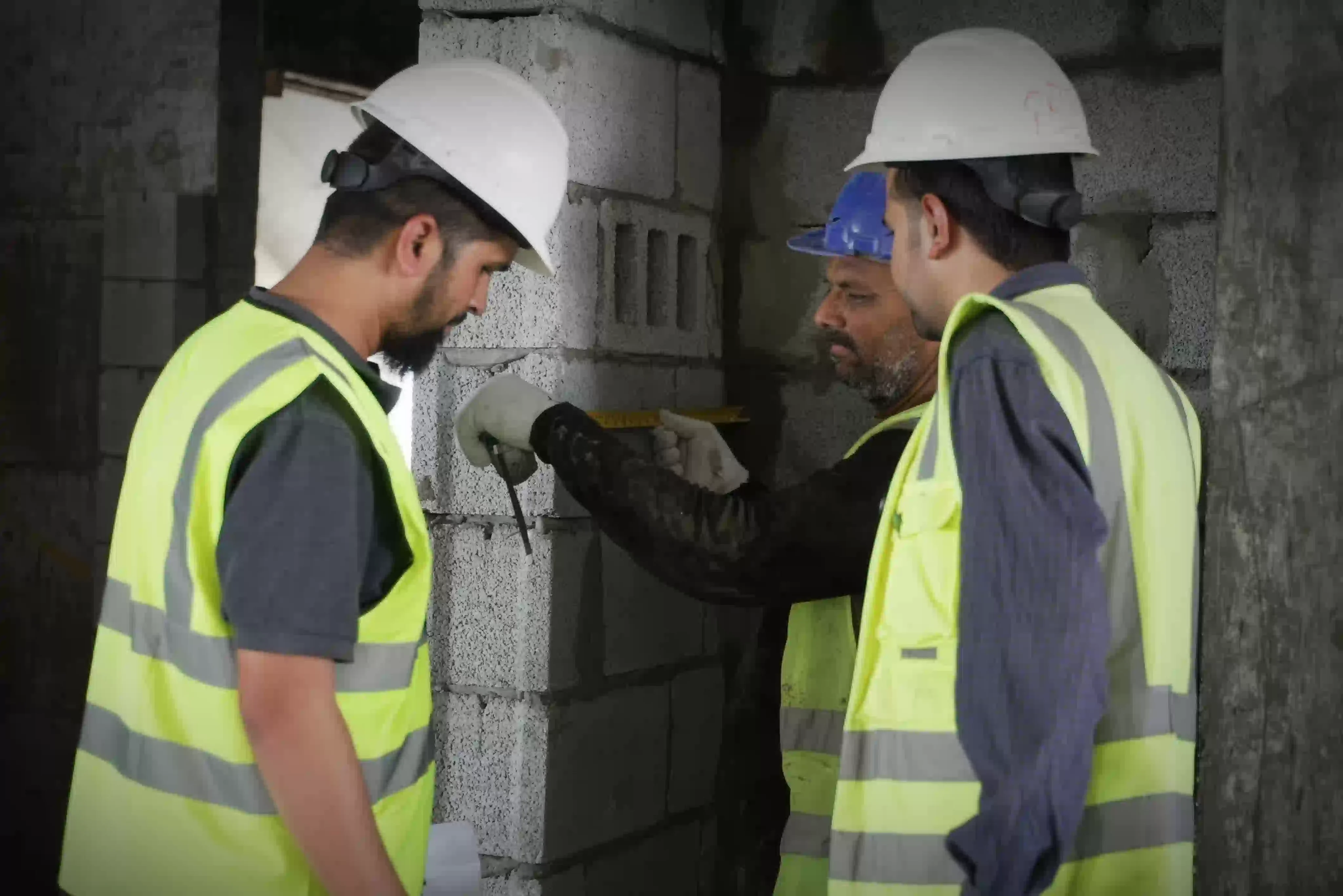 Drei Bauarbeiter mit Schutzhelmen und reflektierenden Westen vermessen eine Betonwand in einem Innenraum.