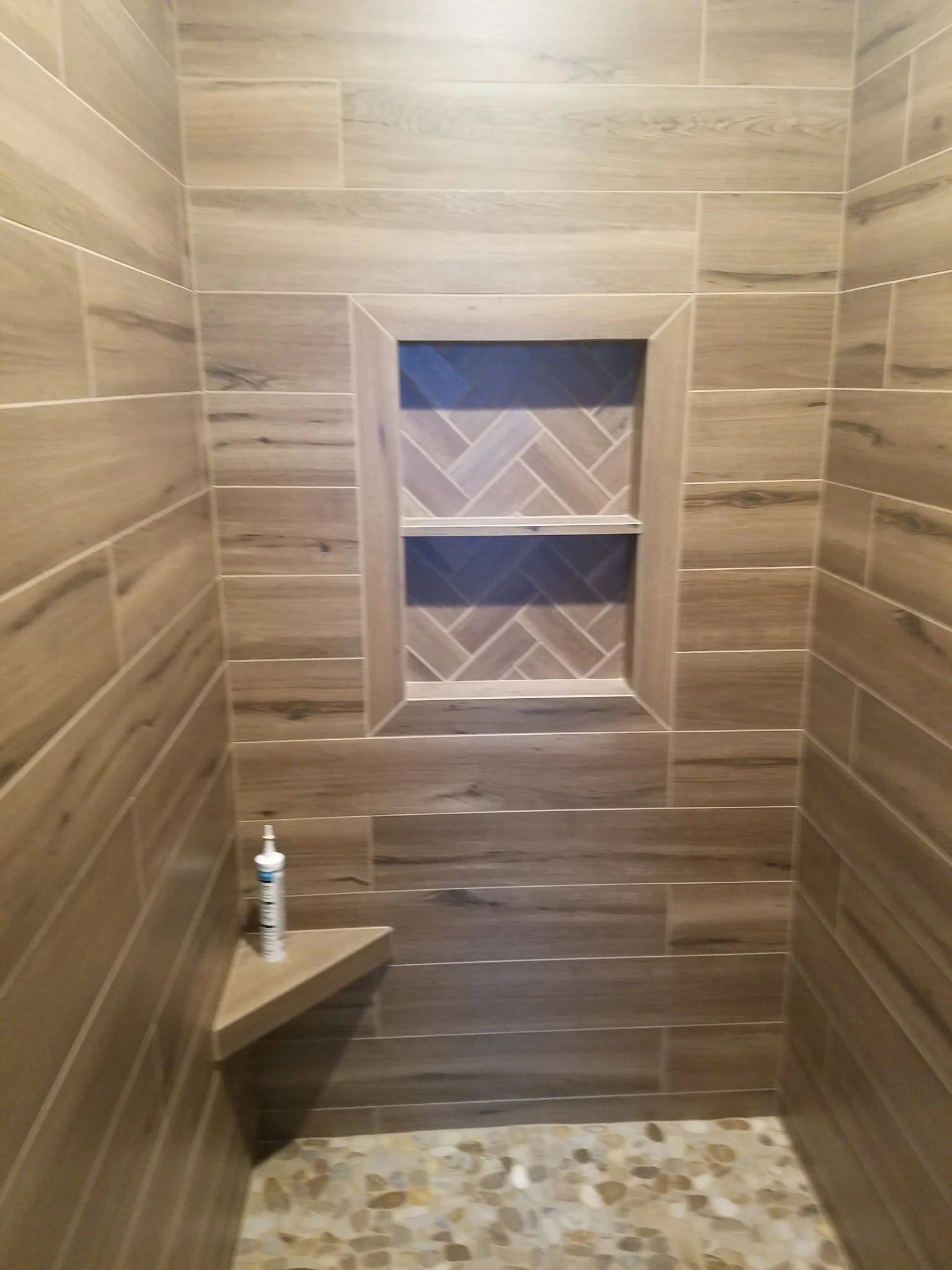 Shower in wood-look plank tile with a framed herringbone niche, corner shelf and pebble floor