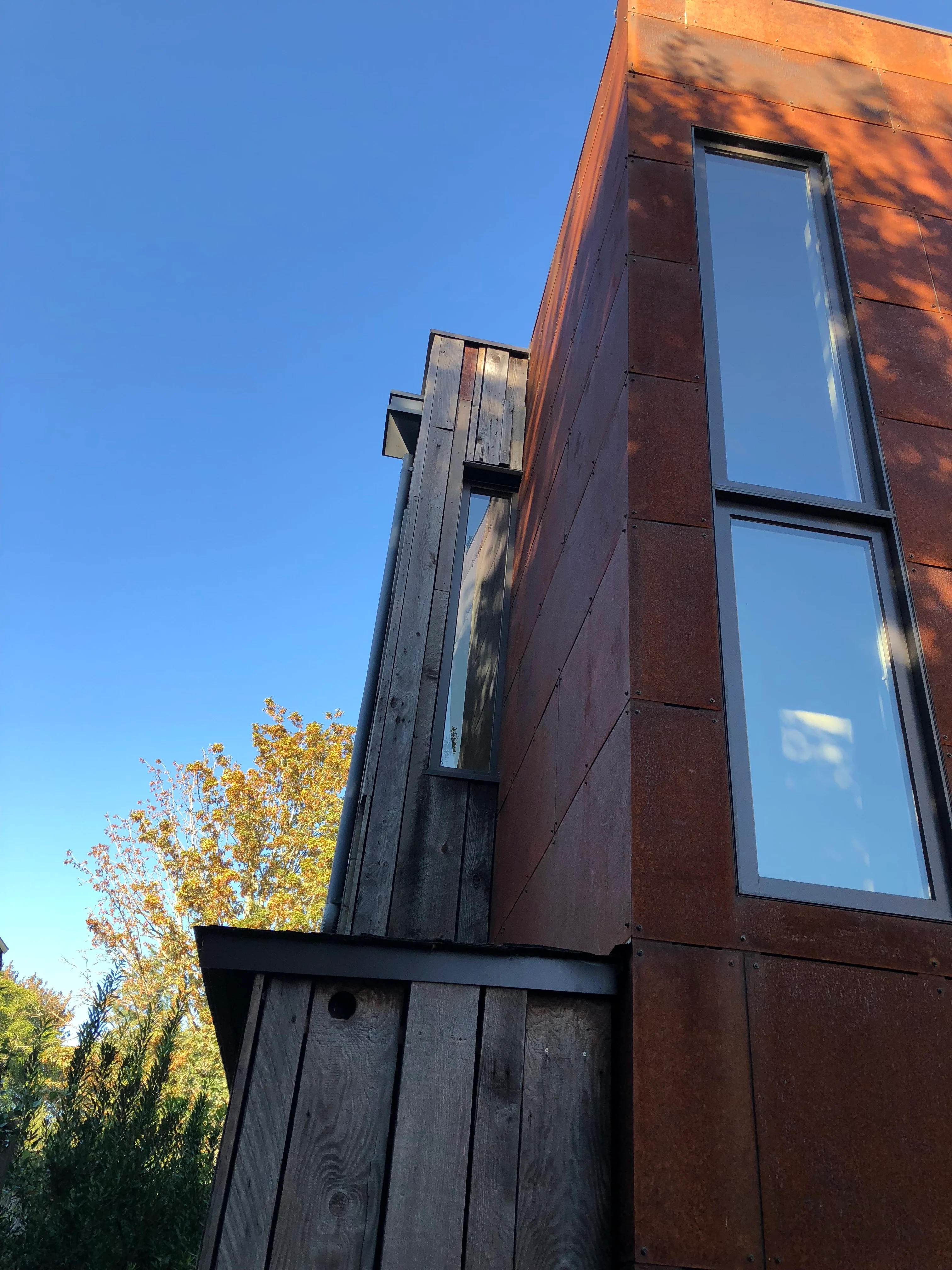 Rust-colored steel panel exterior with tall windows beside weathered wood siding under a clear blue sky