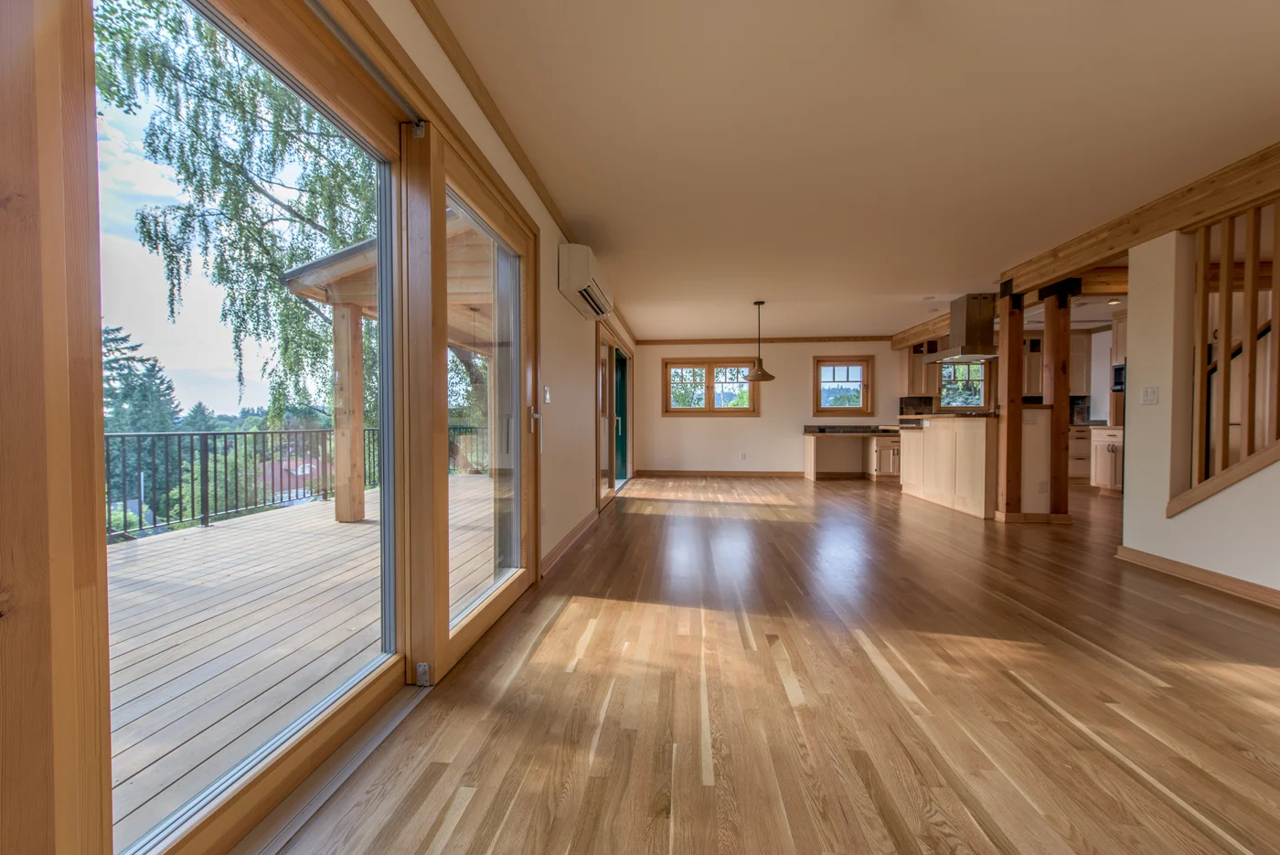 Open living room with light oak floors, a wall of glass doors to a wood deck, and a kitchen beyond