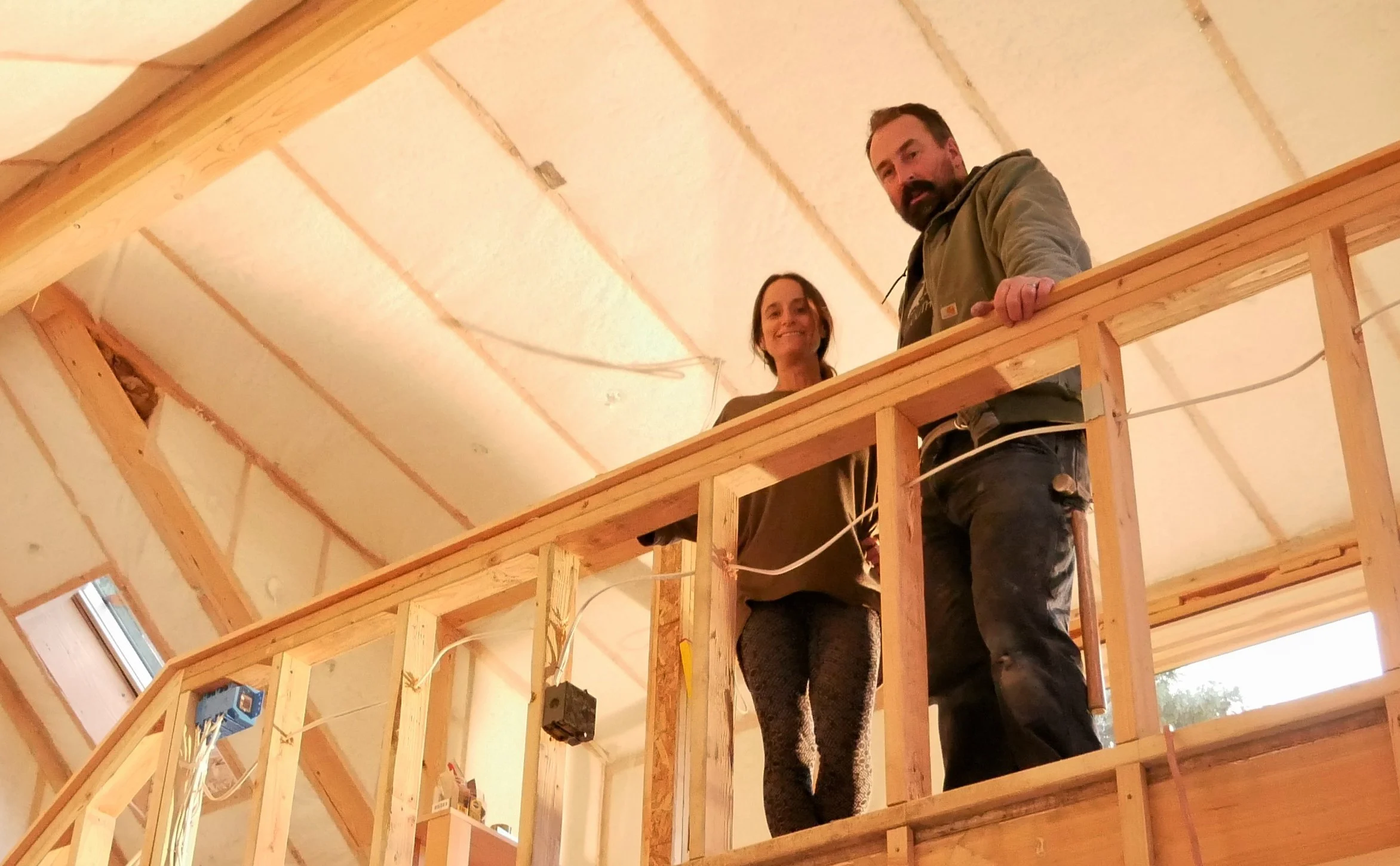 A smiling woman and a bearded man at a wood-framed loft railing under an insulated vaulted ceiling