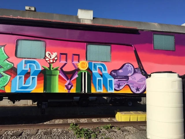 Colorful block-letter graffiti on a pink and orange train car, with a green pipe and a purple cloud shape