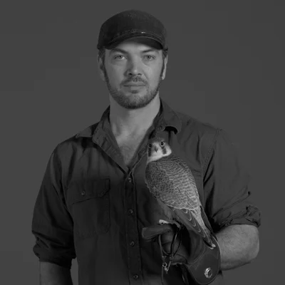Black-and-white portrait of a man in a black cap and dark shirt holding a falcon on a gloved hand against a gray backdrop