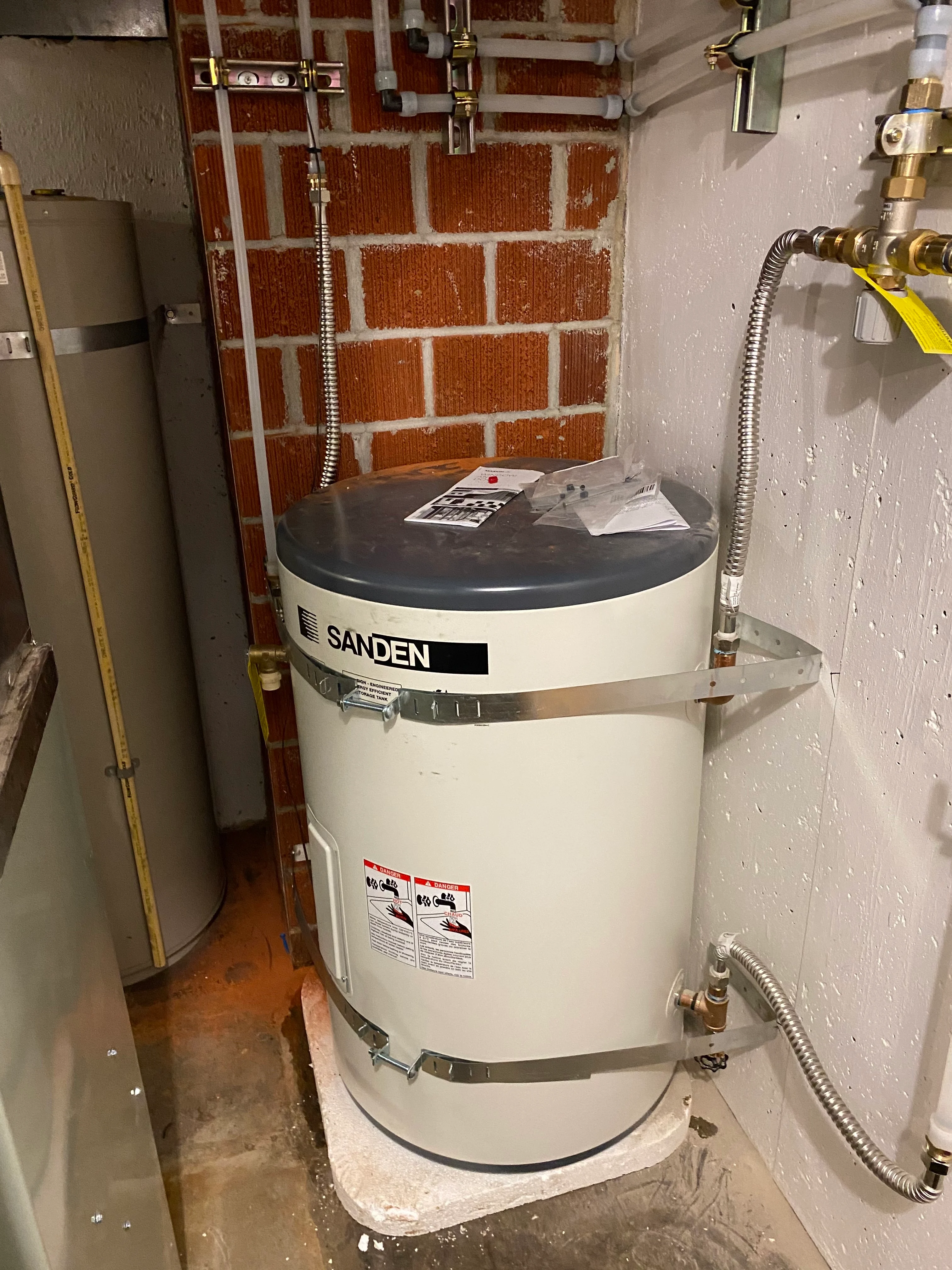 SANDEN water heater in a utility closet beside an exposed brick wall and white plastic water lines