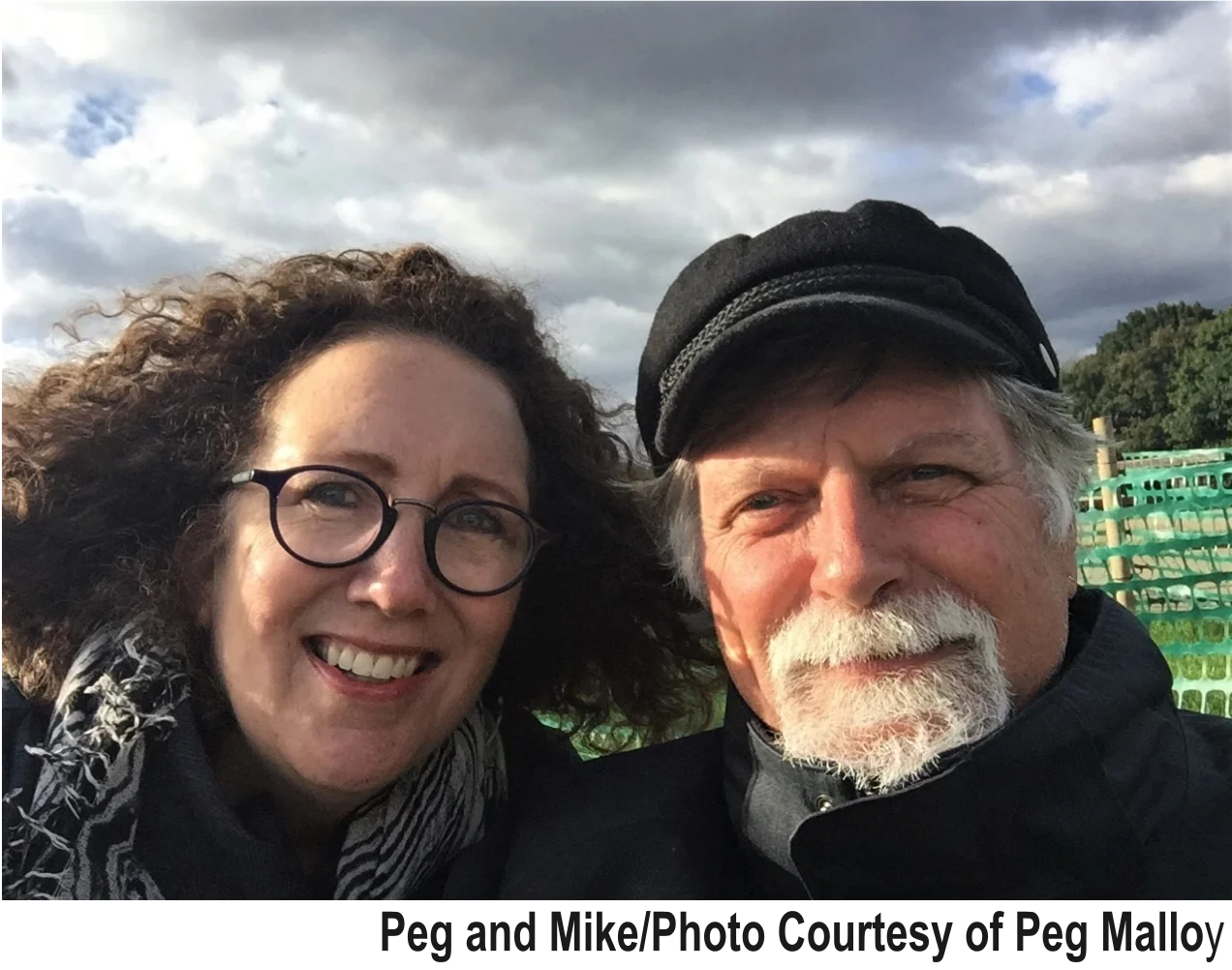 Selfie of a smiling woman with curly hair and round glasses beside a man with a gray beard and black cap, captioned "Peg and Mike"