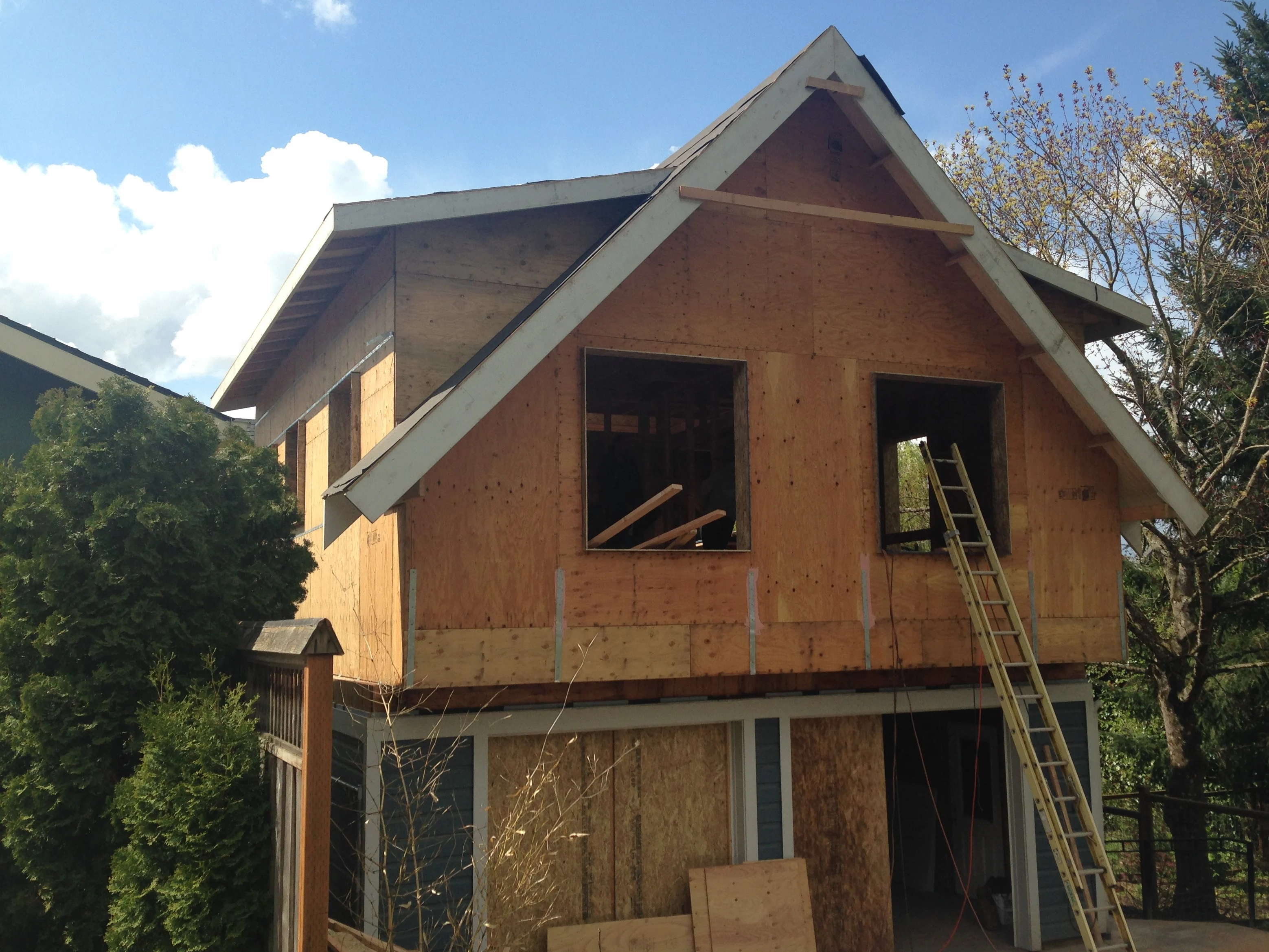Partly sheathed plywood building with a gable roof, open window frames, and a ladder leaning against the wall