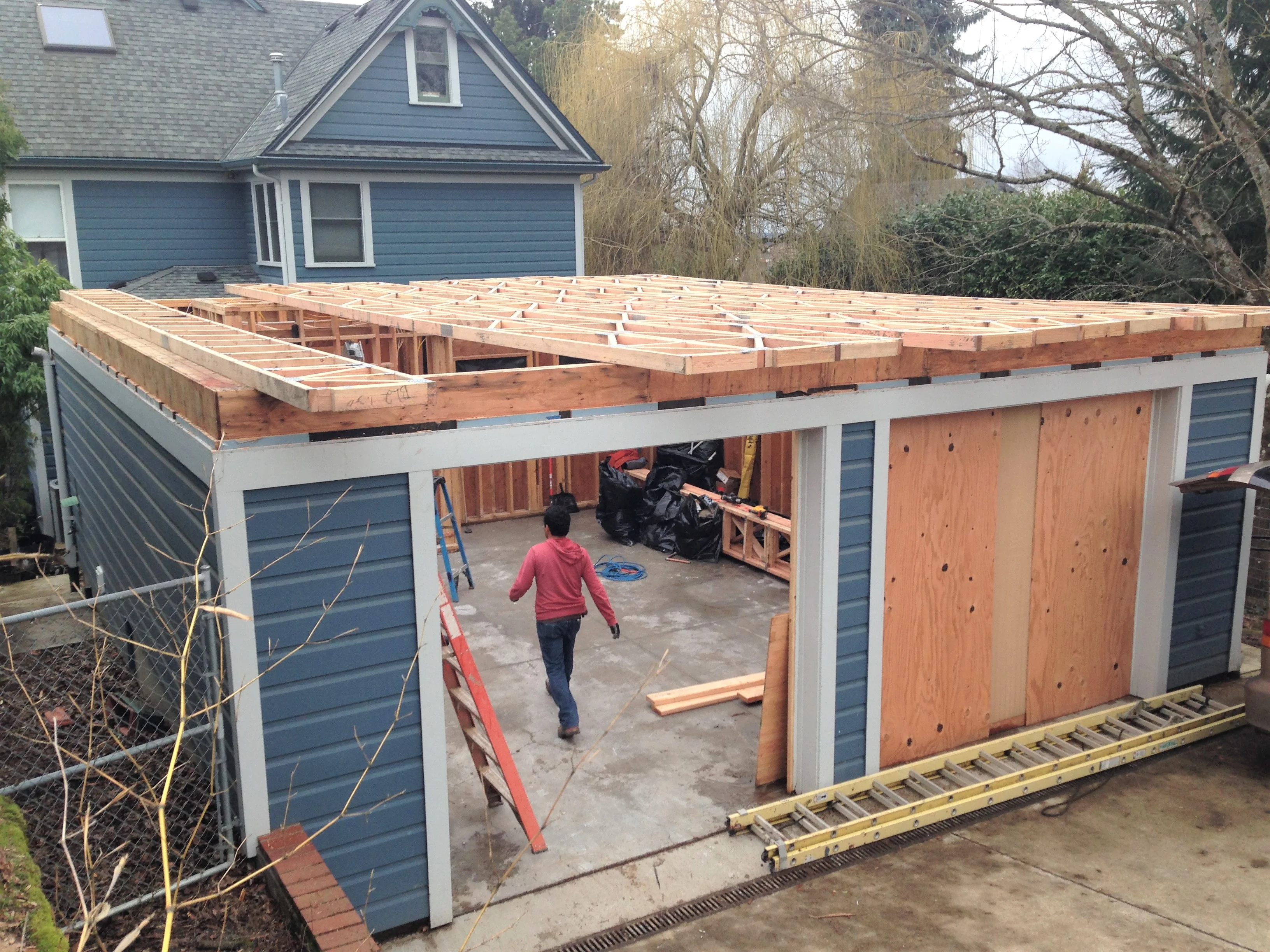 Blue-sided addition under construction with exposed roof framing and a worker in a red hoodie walking inside