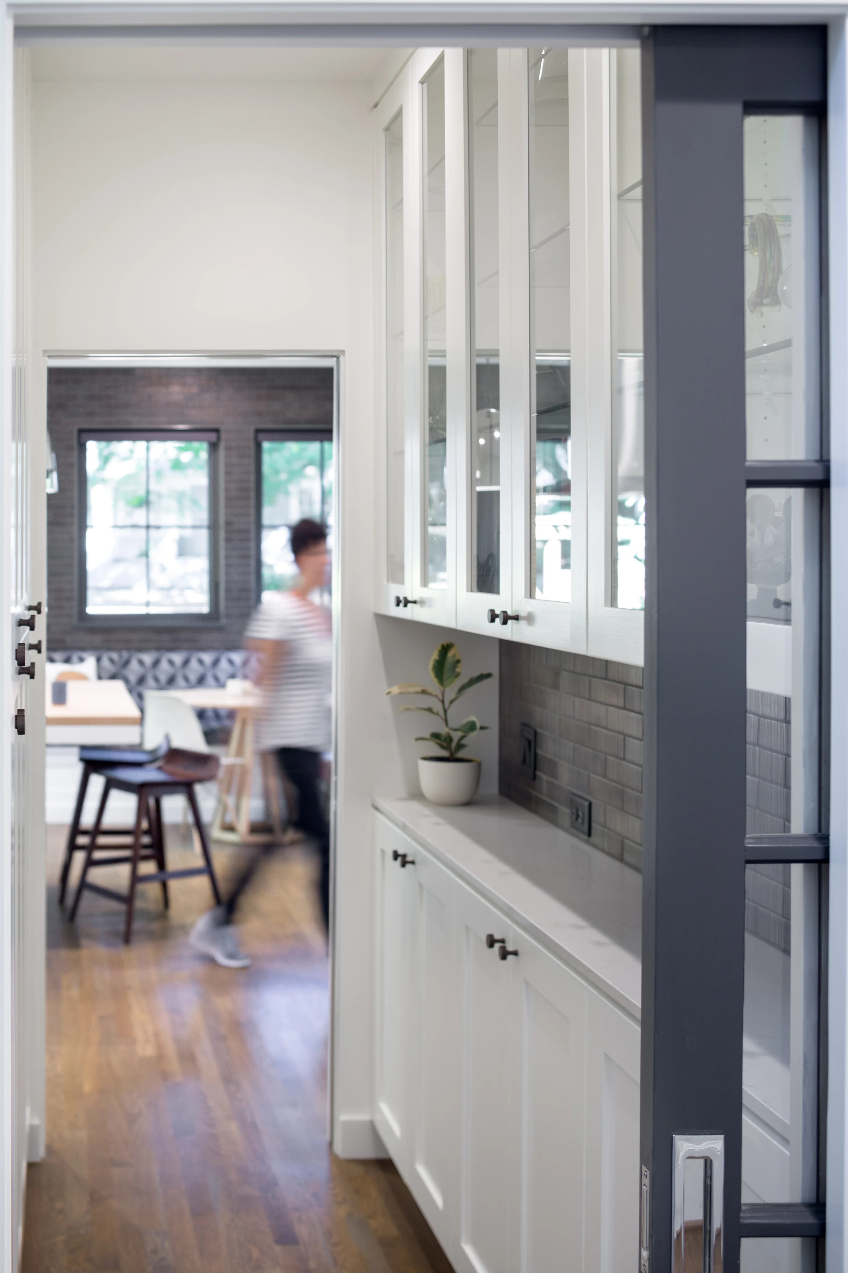 Glass-front white cabinets beside a kitchen doorway, with a blurred person walking past stools and a potted plant on the counter