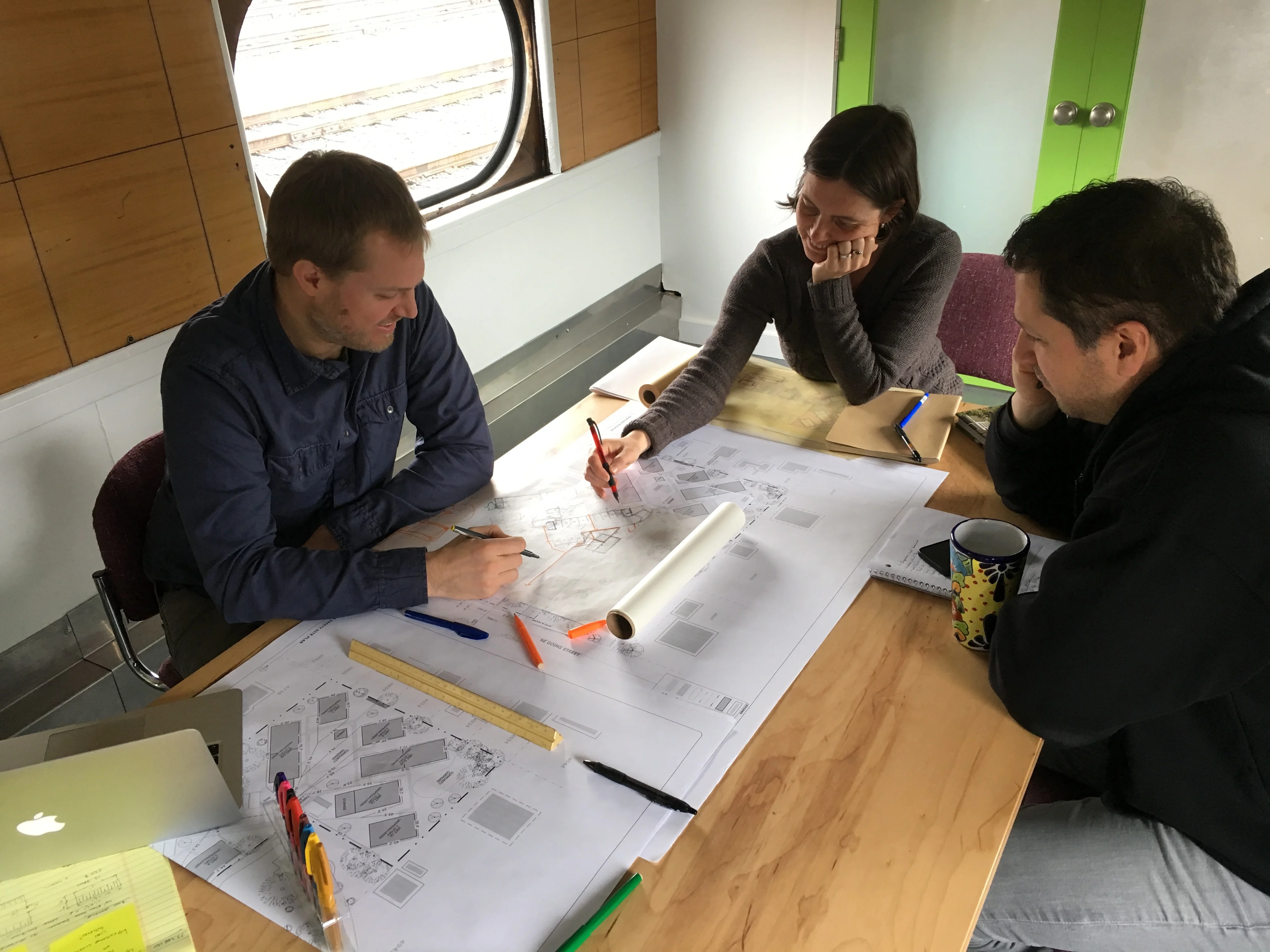 Three people reviewing site plan drawings on a wooden table inside a railcar, with pens and a rolled sheet of paper