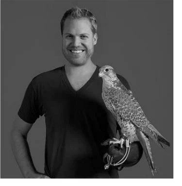 Jonathan Lundstrom, architect, smiling in a dark V-neck shirt and holding a falcon on a gloved hand against a gray backdrop