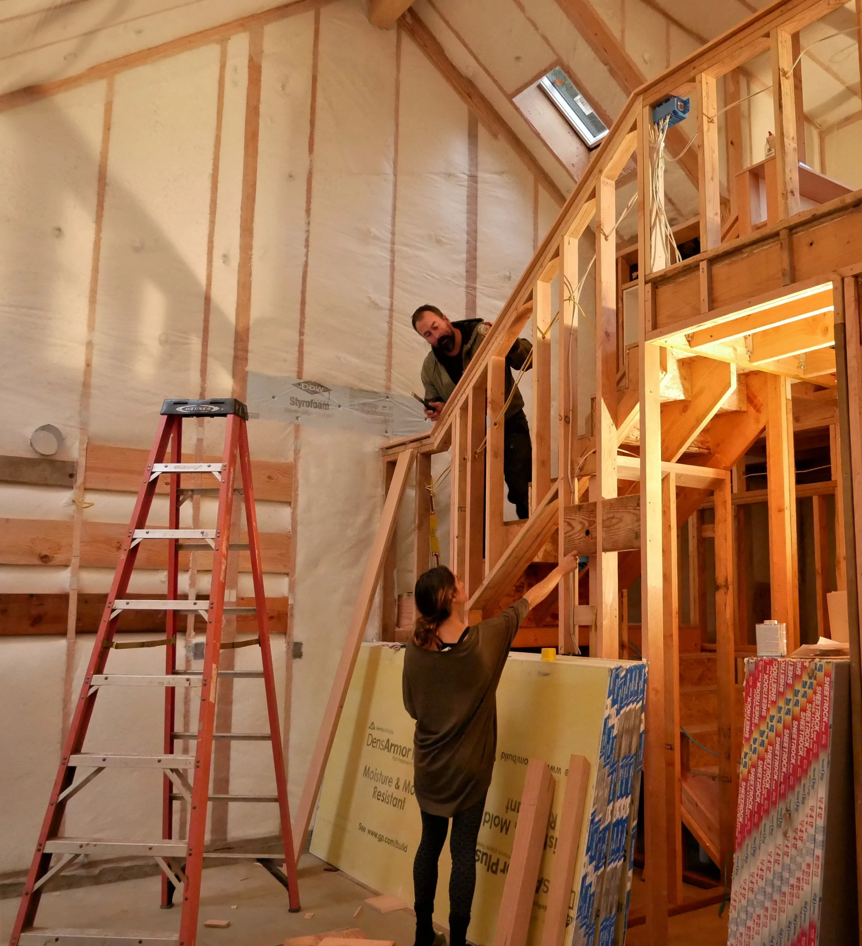 A woman reaching up to a bearded man on a framed wood staircase in an ADU under construction, with insulated walls and a red ladder