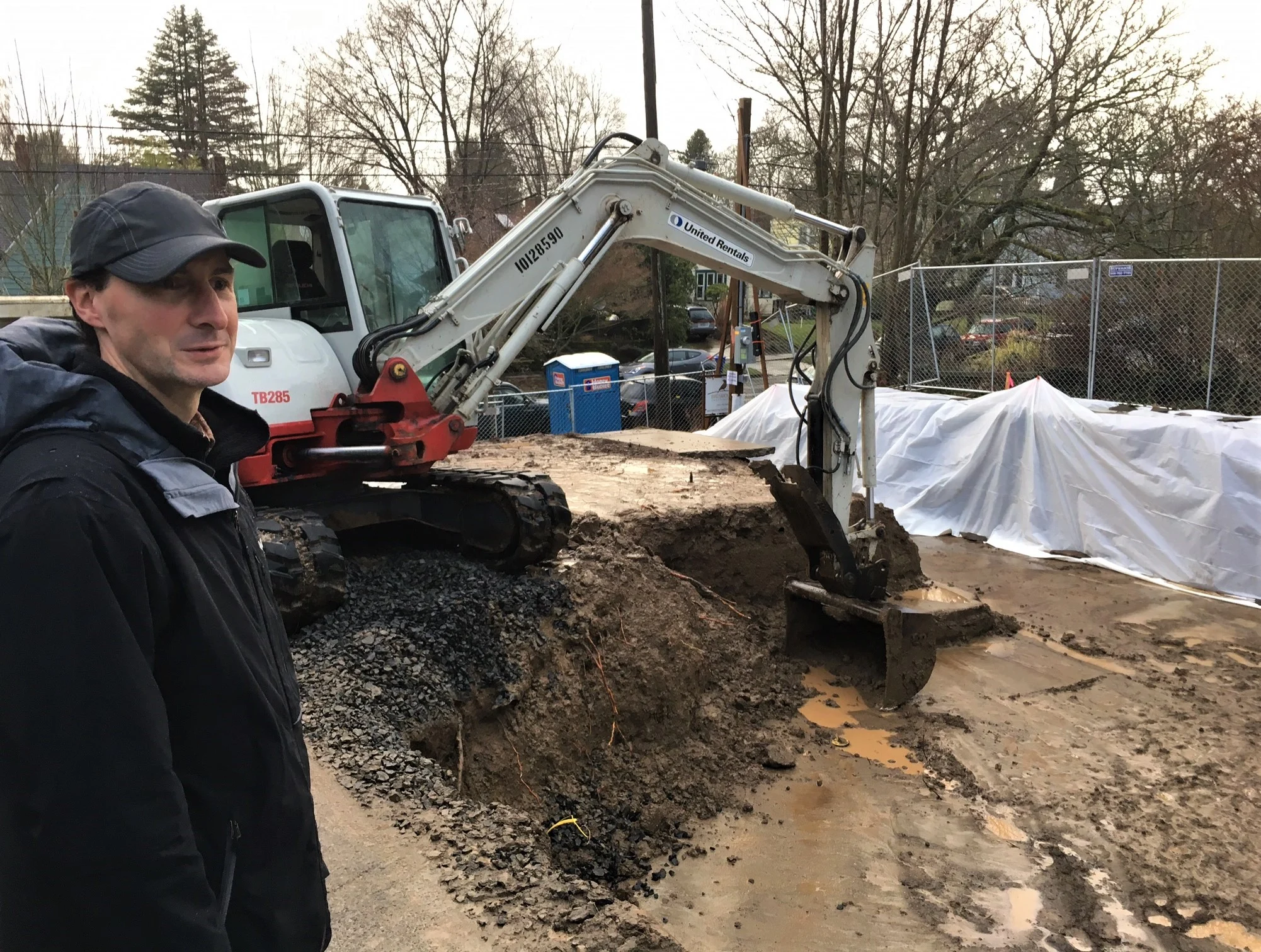 Man in a dark jacket and cap beside a United Rentals excavator at a muddy construction site with a chain-link fence