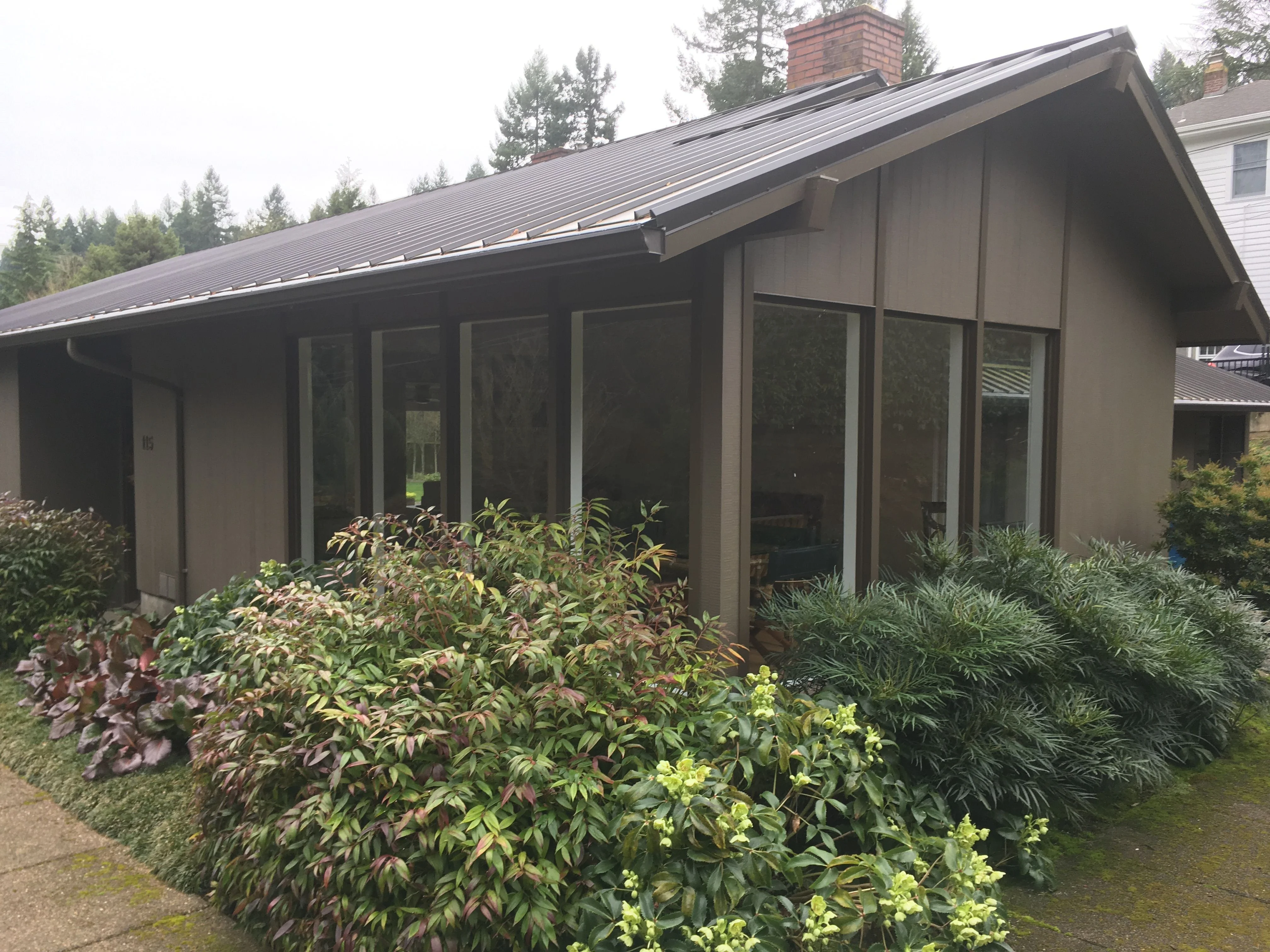 Single-story brown house with a dark metal roof, a brick chimney, and large windows framed by dense shrubs