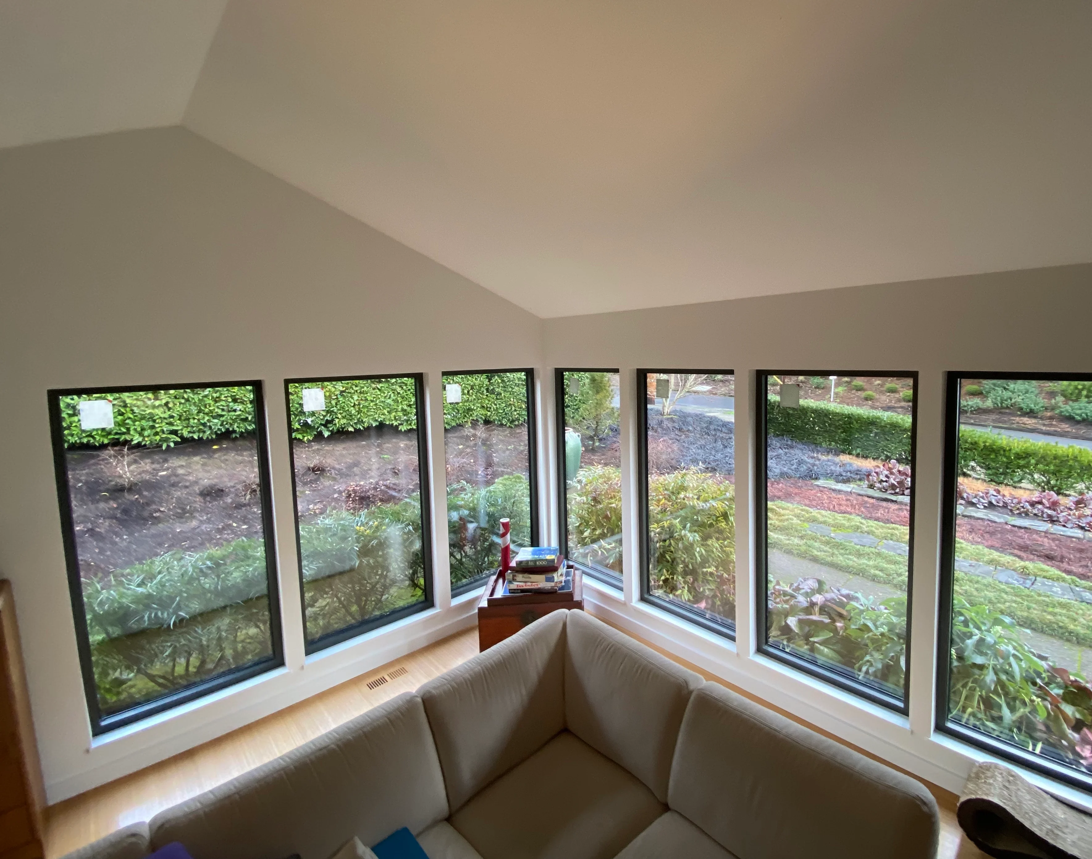 Living room with a vaulted ceiling, a row of black-framed windows overlooking a garden, and a beige sectional sofa