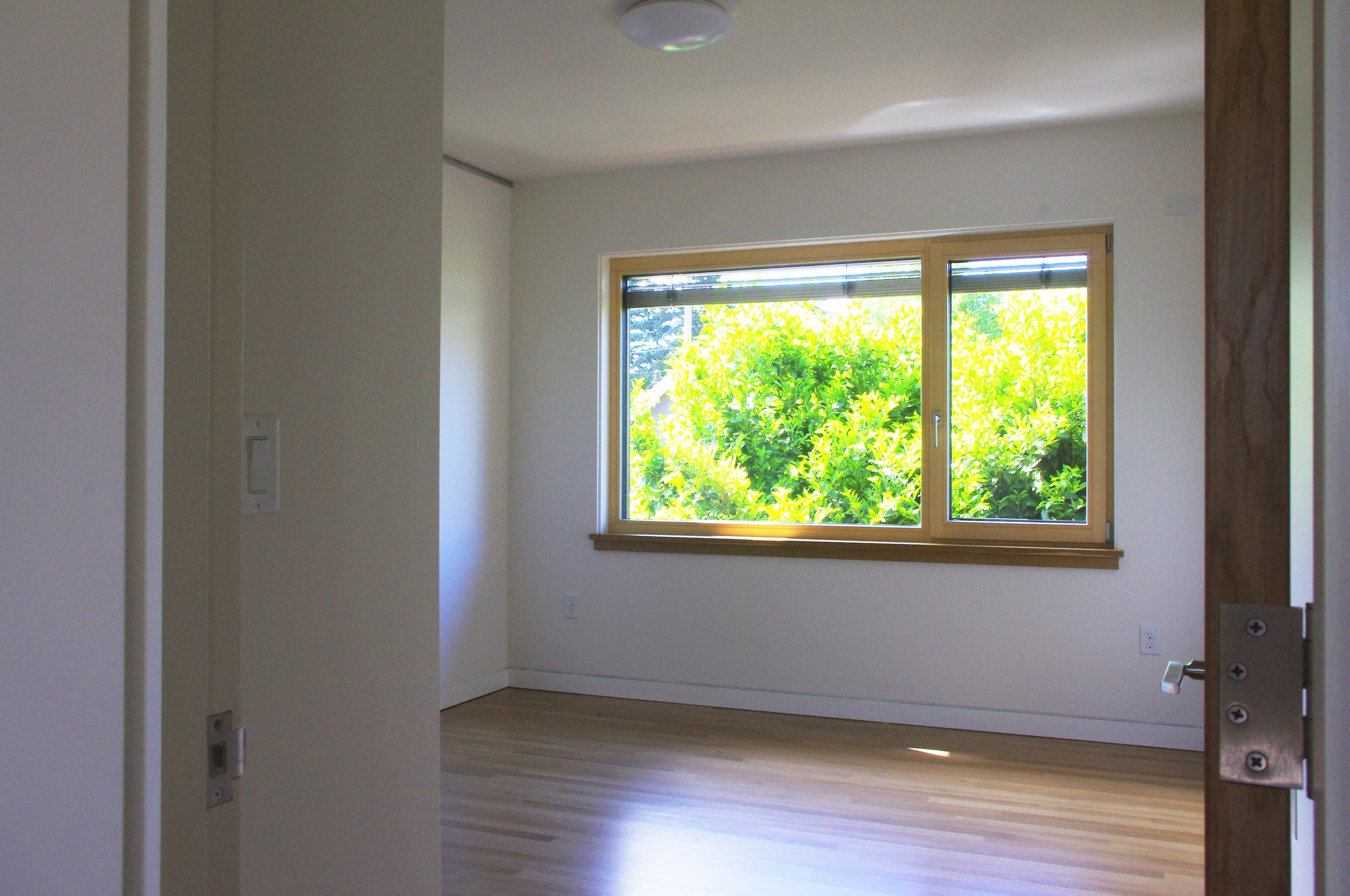 Empty bedroom seen through an open doorway, with a wood-framed window looking onto green shrubs and light oak floors