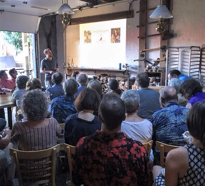 Presenter in a gray shirt speaking to a seated audience in a brick-walled room, with a slide projected behind him