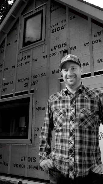 Josh Salinger smiling in a plaid shirt and cap in front of a house wall wrapped in SIGA sheathing
