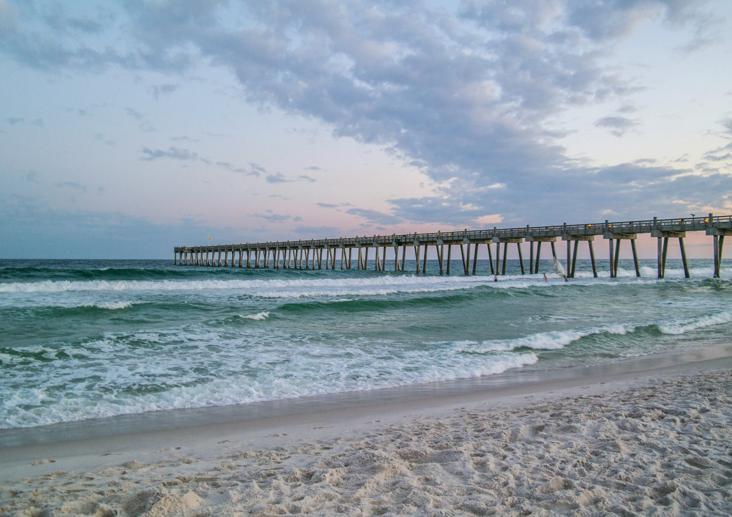 Long wooden fishing pier with lights extends over foamy ocean waves beneath a cloudy blue evening sky.