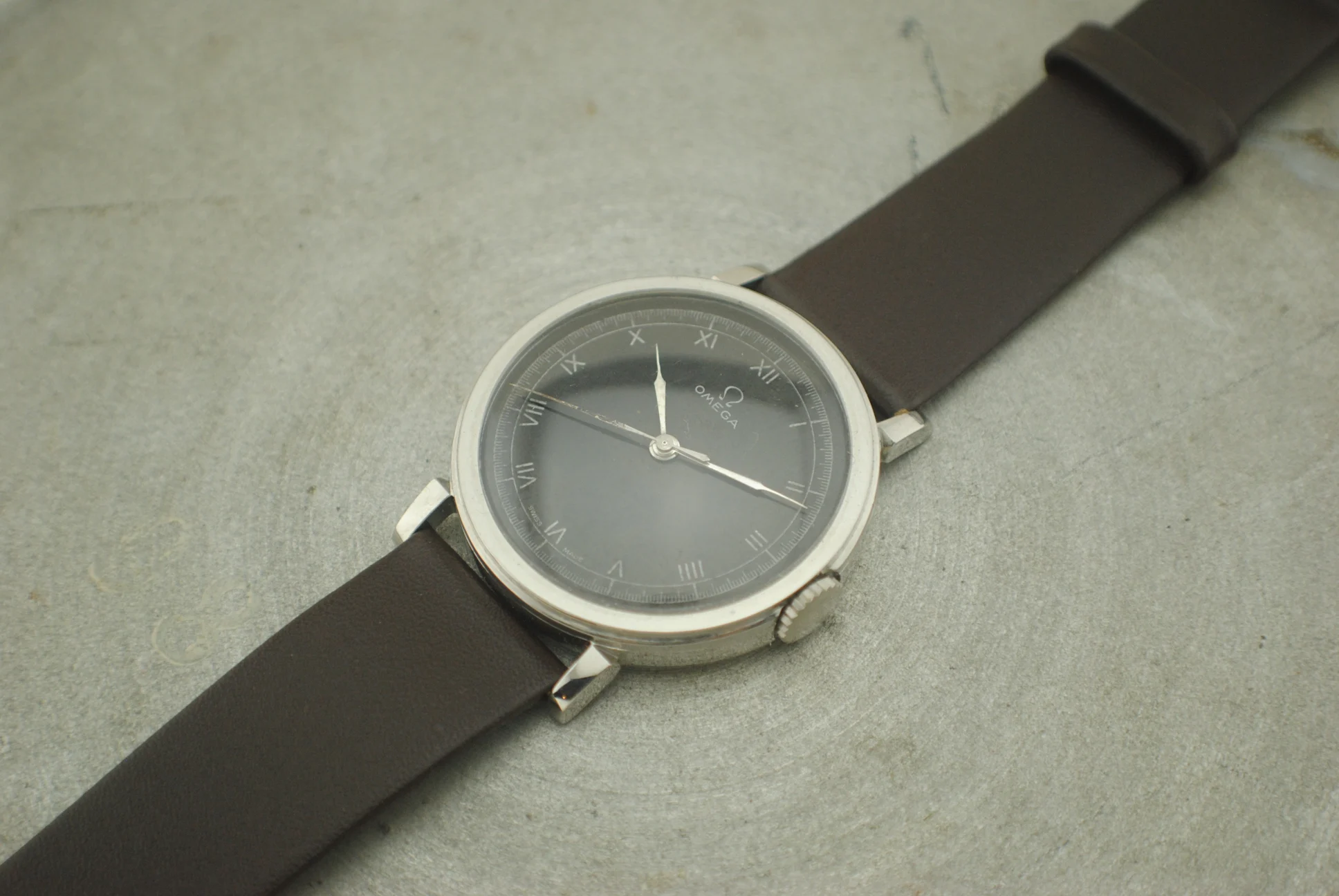Crystal-on front view of the Omega CK2015 Fagiolino with black Roman-numeral dial and brown leather strap on a grey surface