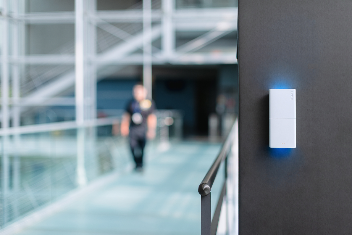 A wall-mounted Firefly device with a glowing indicator on a dark panel, with a security responder approaching down a glass-enclosed corridor in soft focus.