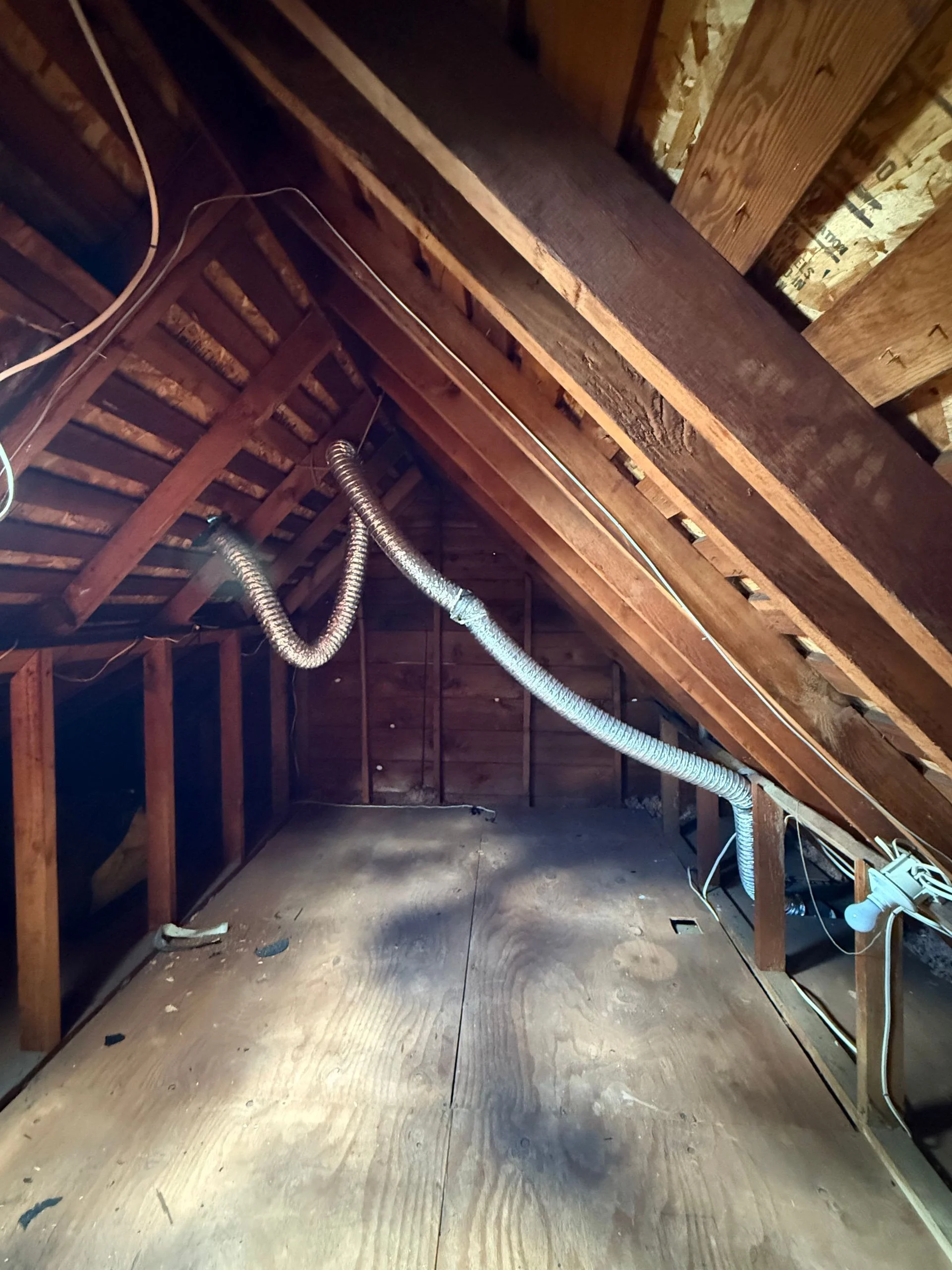 An unfinished attic with wooden rafters and floorboards, featuring a flexible metallic ventilation duct suspended across.
