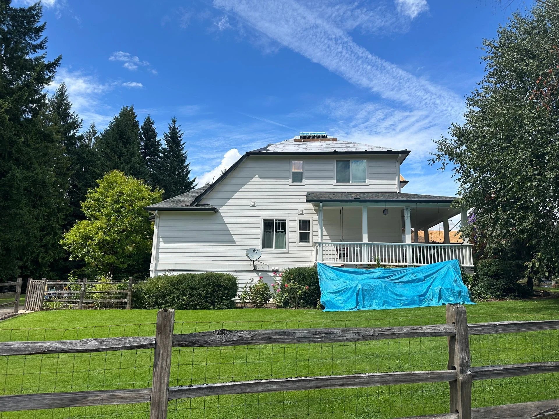 A white two-story house with a wooden porch and blue tarp stands in a green yard behind a rustic wooden fence.