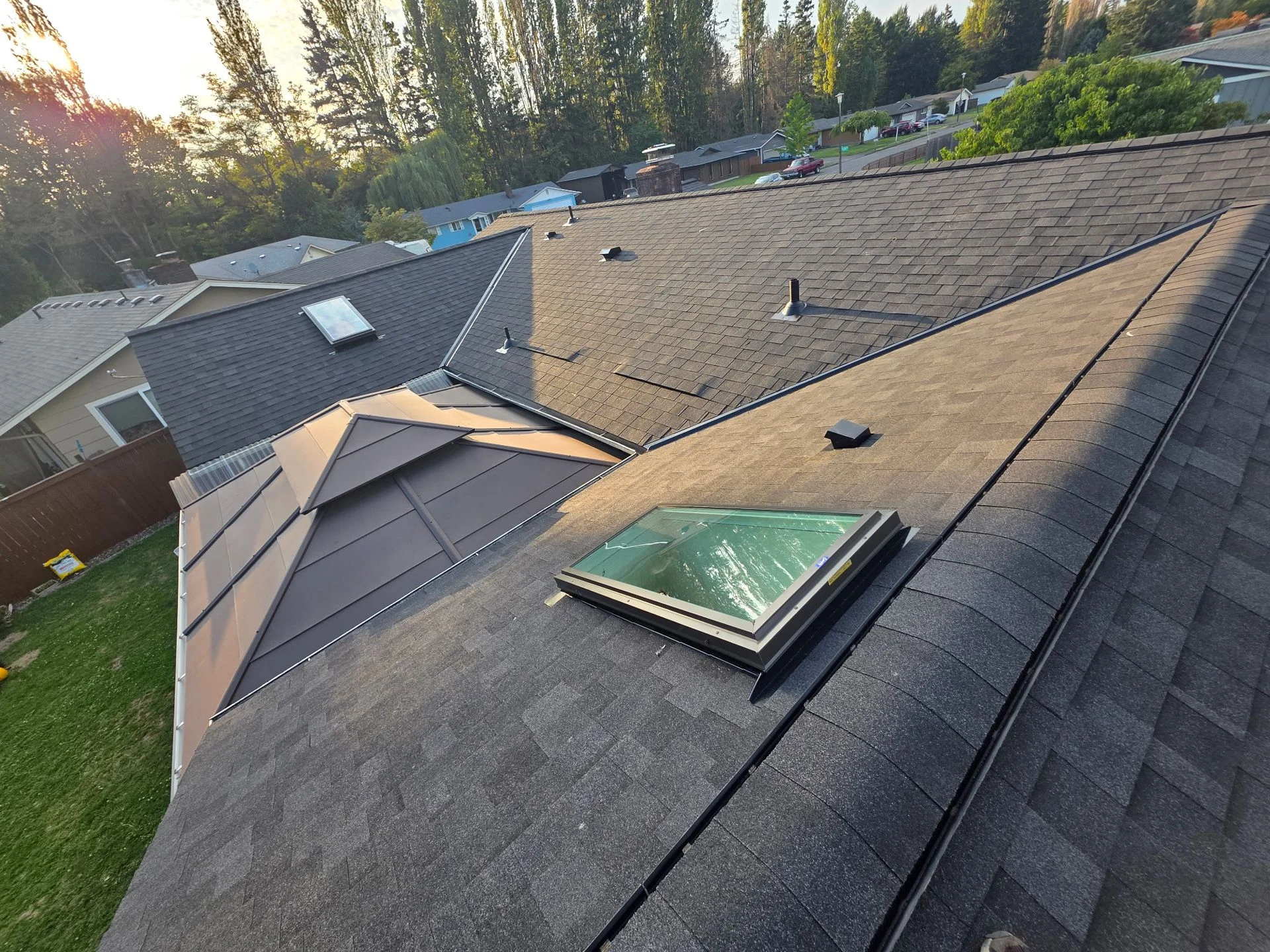 A high-angle view of a residential shingled roof with a skylight, a roof vent, and a metal-roofed addition below.