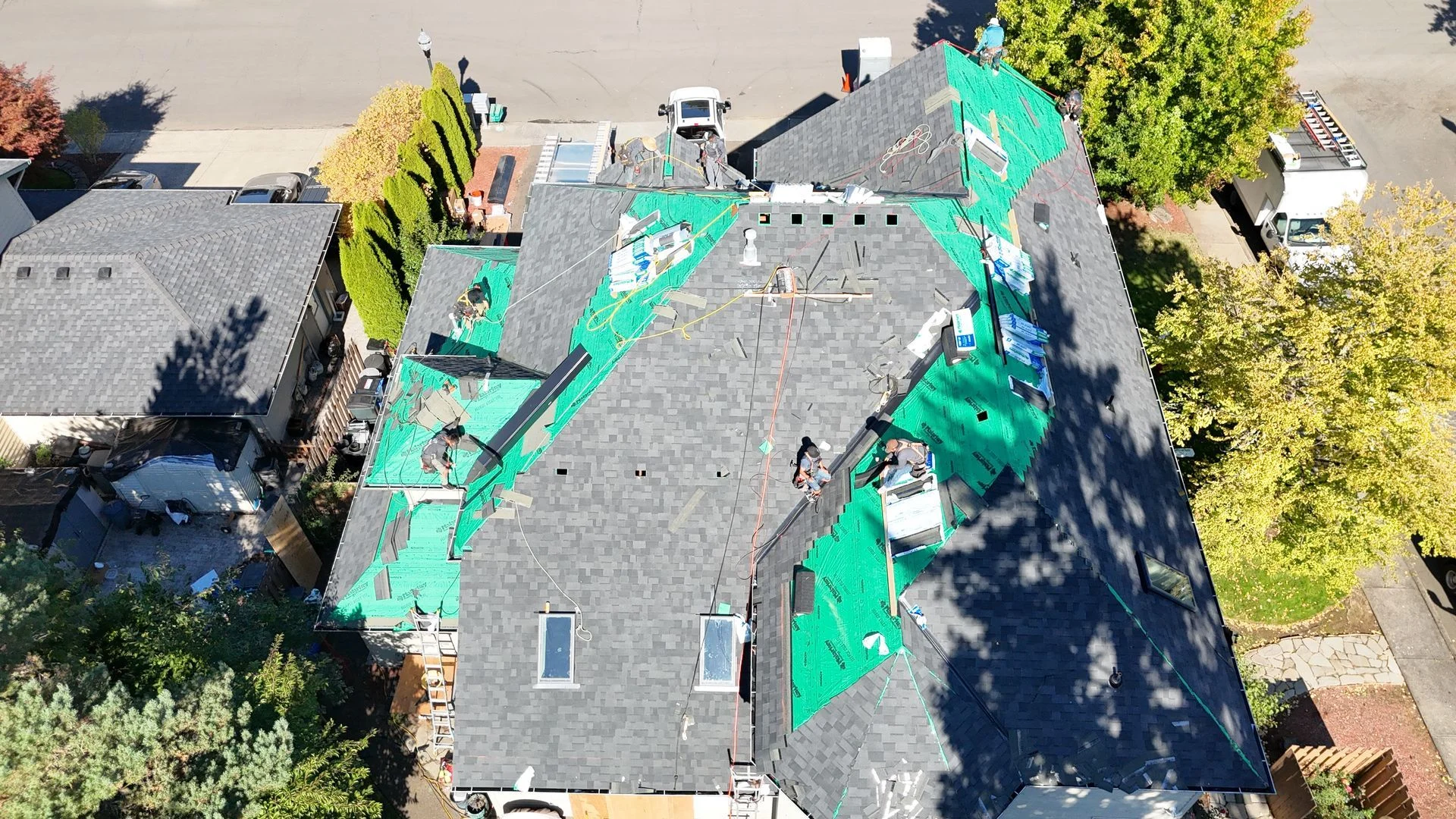 An aerial view of a residential roof undergoing construction, showing shingles partially removed to reveal green underlay.