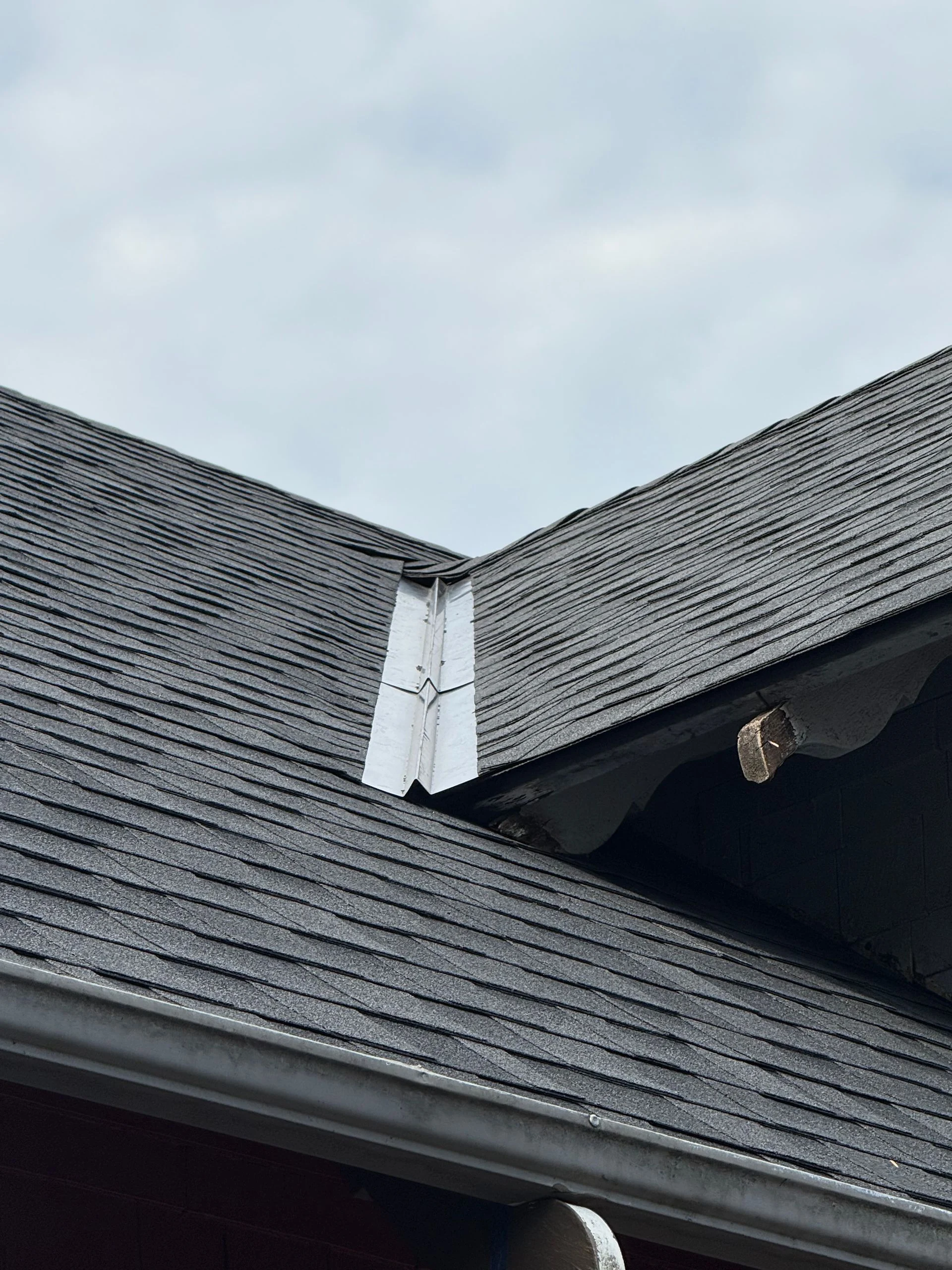 A close-up of a roof valley with grey shingles and a metal flashing strip against a cloudy sky.