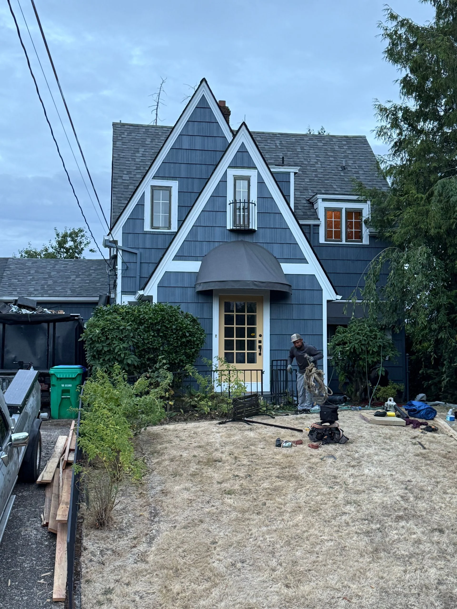 A two-story blue Tudor-style house with a front door awning and a worker in the yard under an overcast sky.