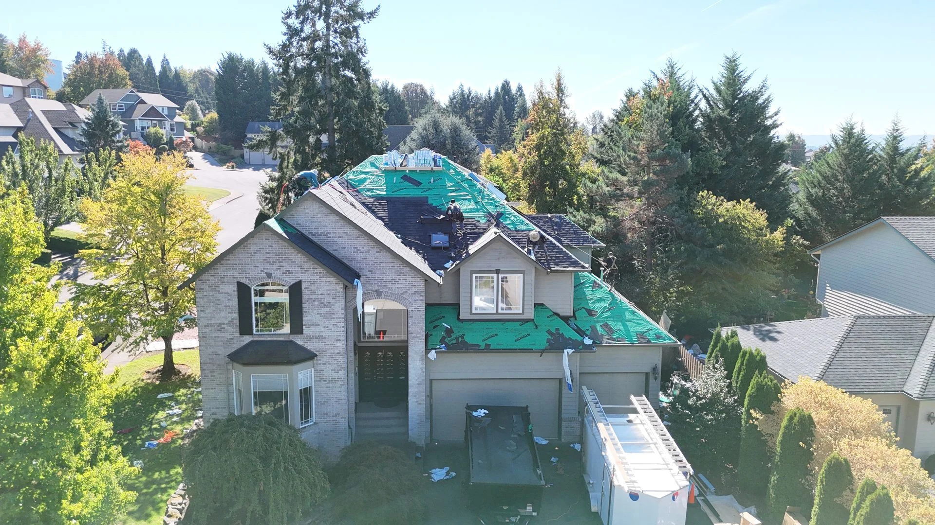A suburban house under renovation, featuring a partially re-roofed structure with green underlayment and a trailer nearby.