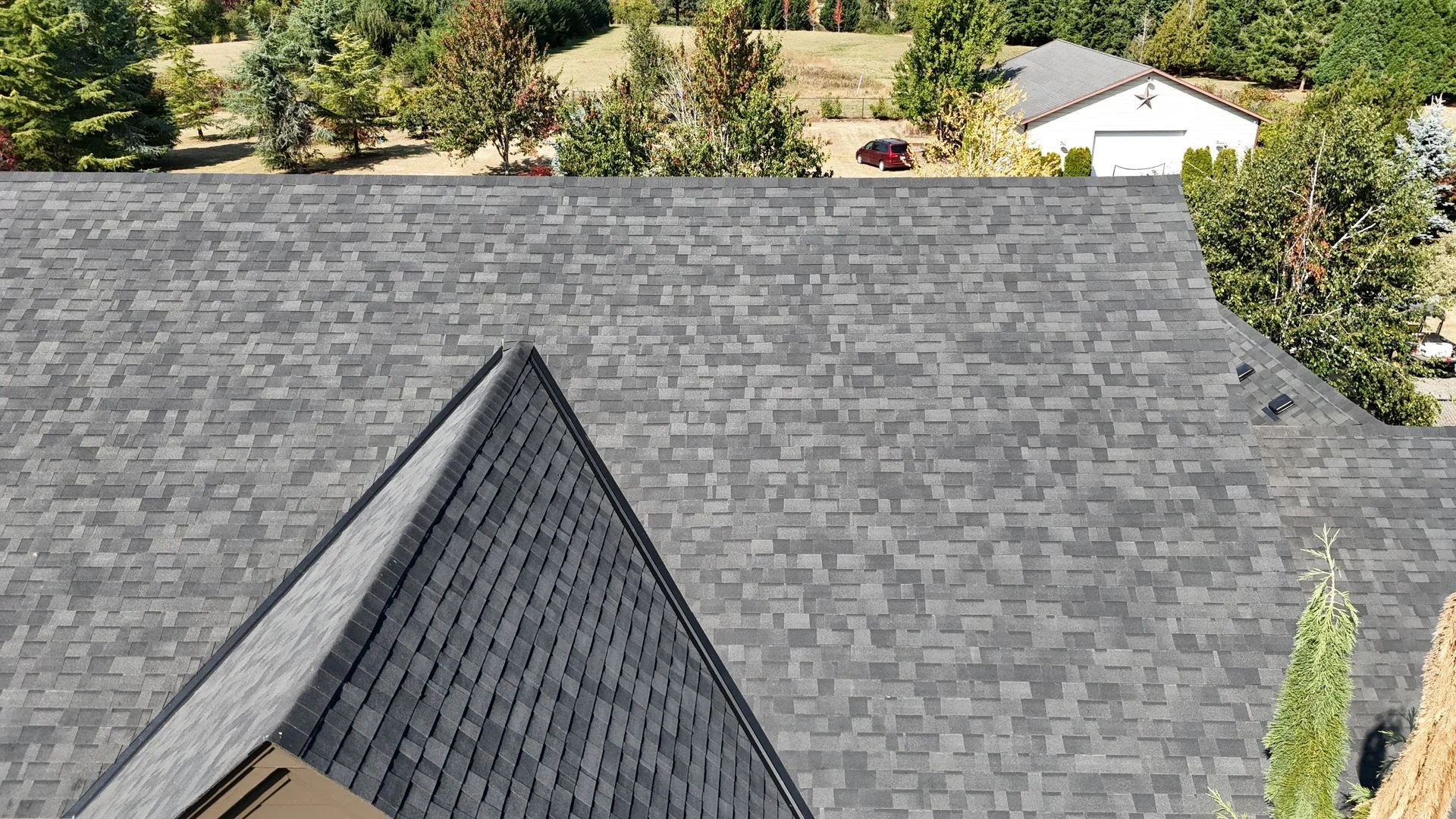 A high-angle view of a grey architectural asphalt shingle roof on a house with trees and a detached garage in the distance.