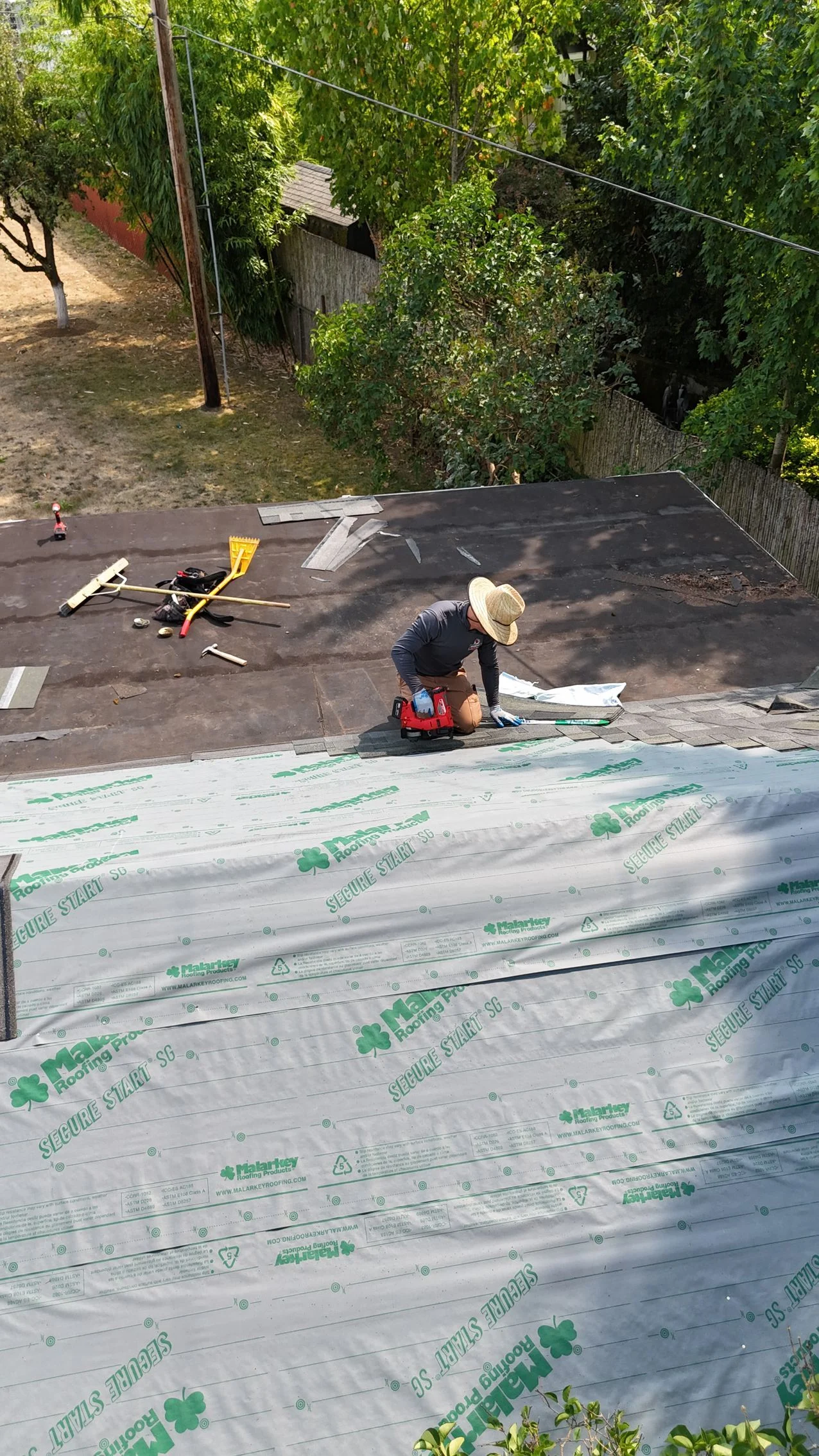 A person wearing a sun hat works on a residential roof, installing gray underlayment over a section of dark shingles.