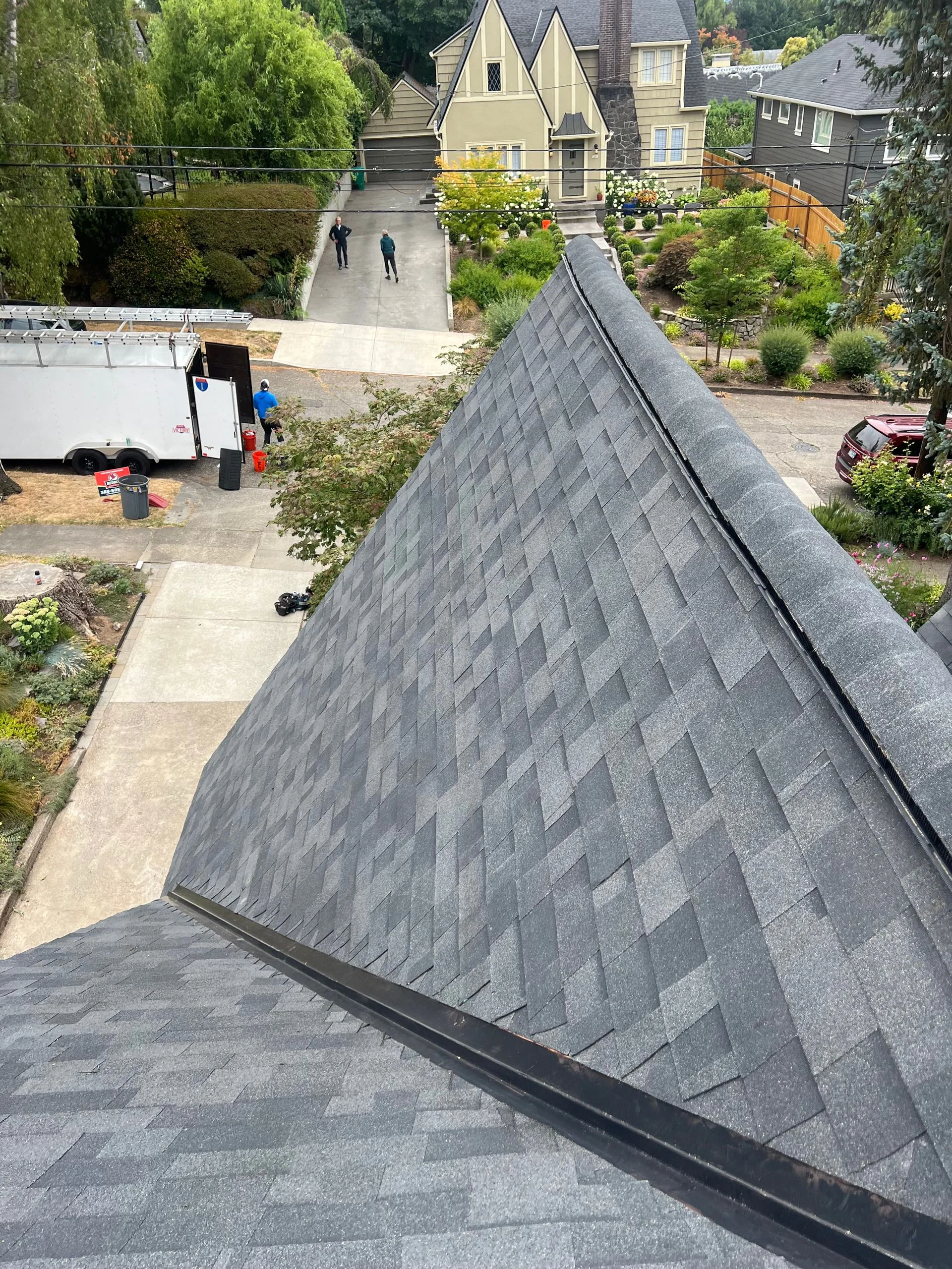 A high-angle view looking down at a newly shingled gray roof, with a construction trailer and house visible below.
