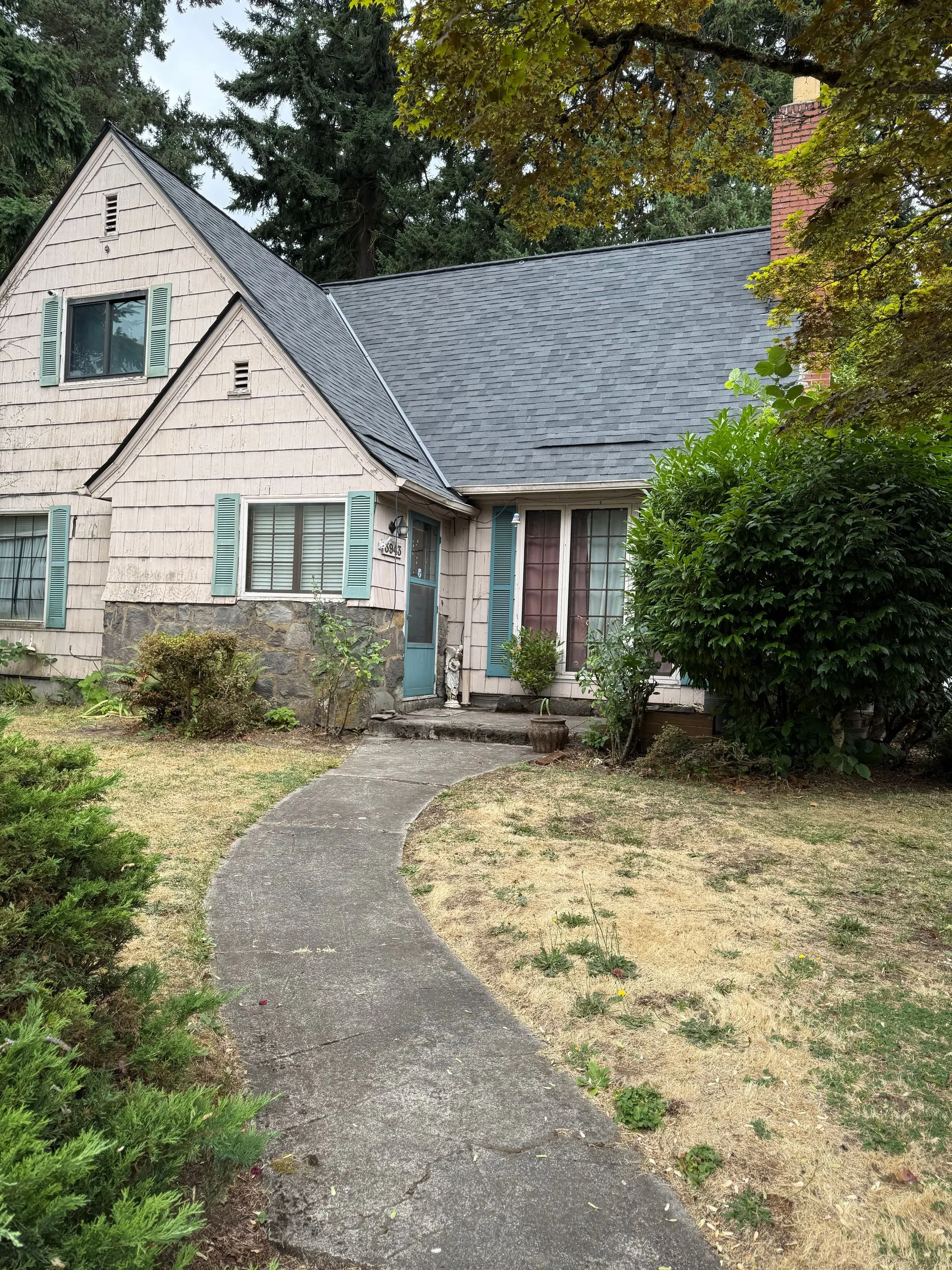 A two-story pink Tudor-style house with a gray roof and stone foundation sits behind a concrete path and dry lawn.